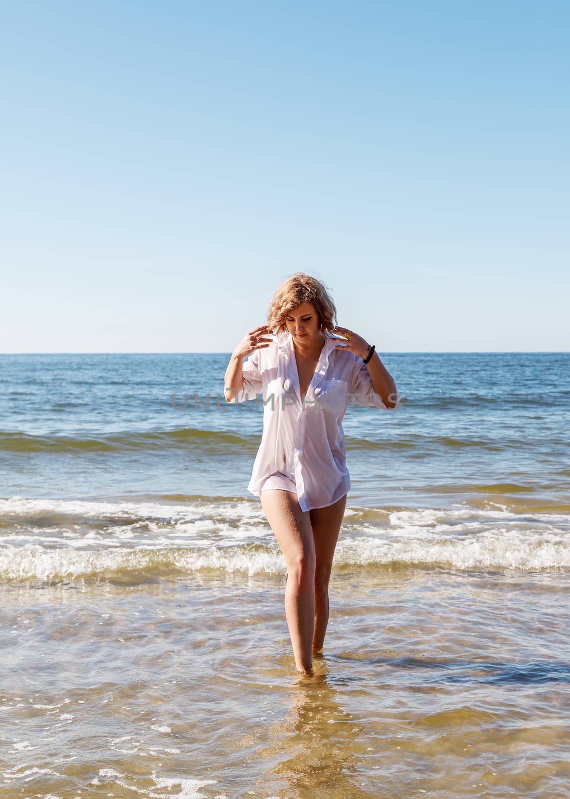 young blonde woman in a wet white shirt coming out of the water near the seashore on sunny summer day
