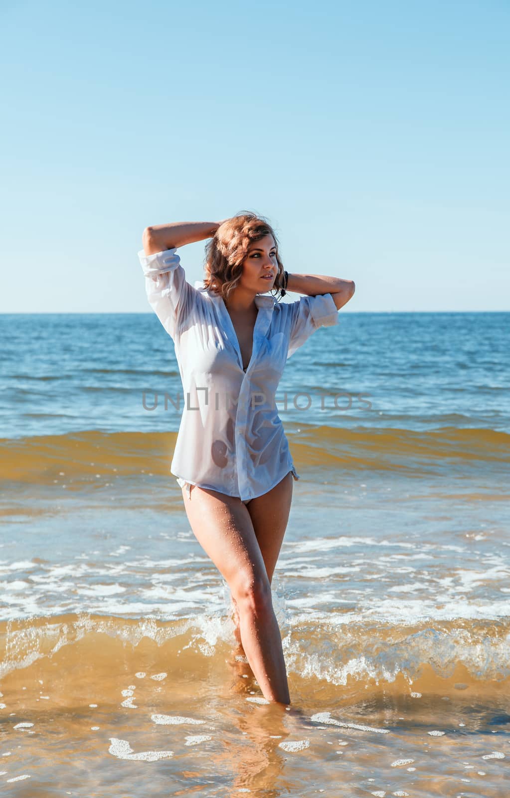 young blonde woman in a wet white shirt standing in the water near the seashore with her hands behind her head on sunny summer day