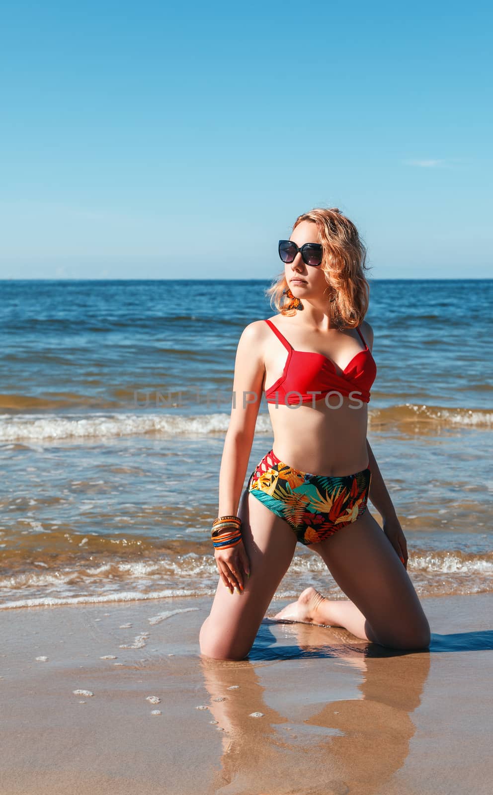 young blonde girl in a red swimsuit and sunglasses standing on the seashore on sunny summer day