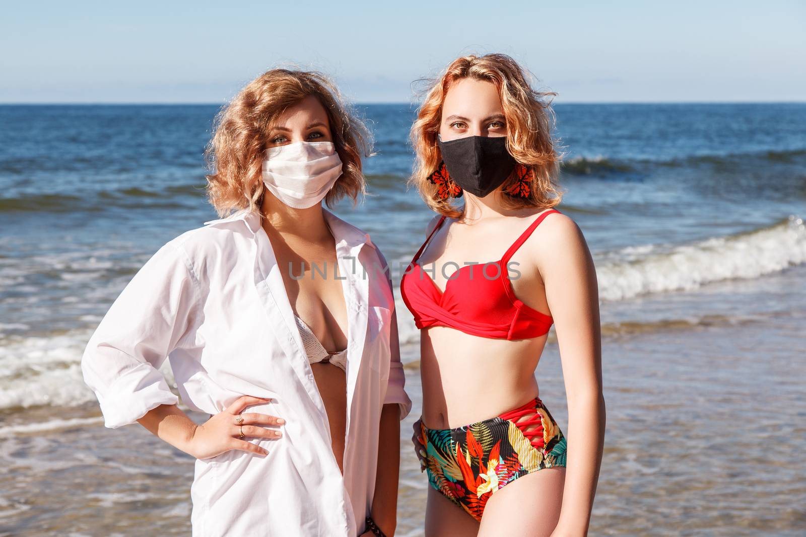two young blonde girls in a swimsuits and protective masks standing on the seashore on sunny summer day closeup