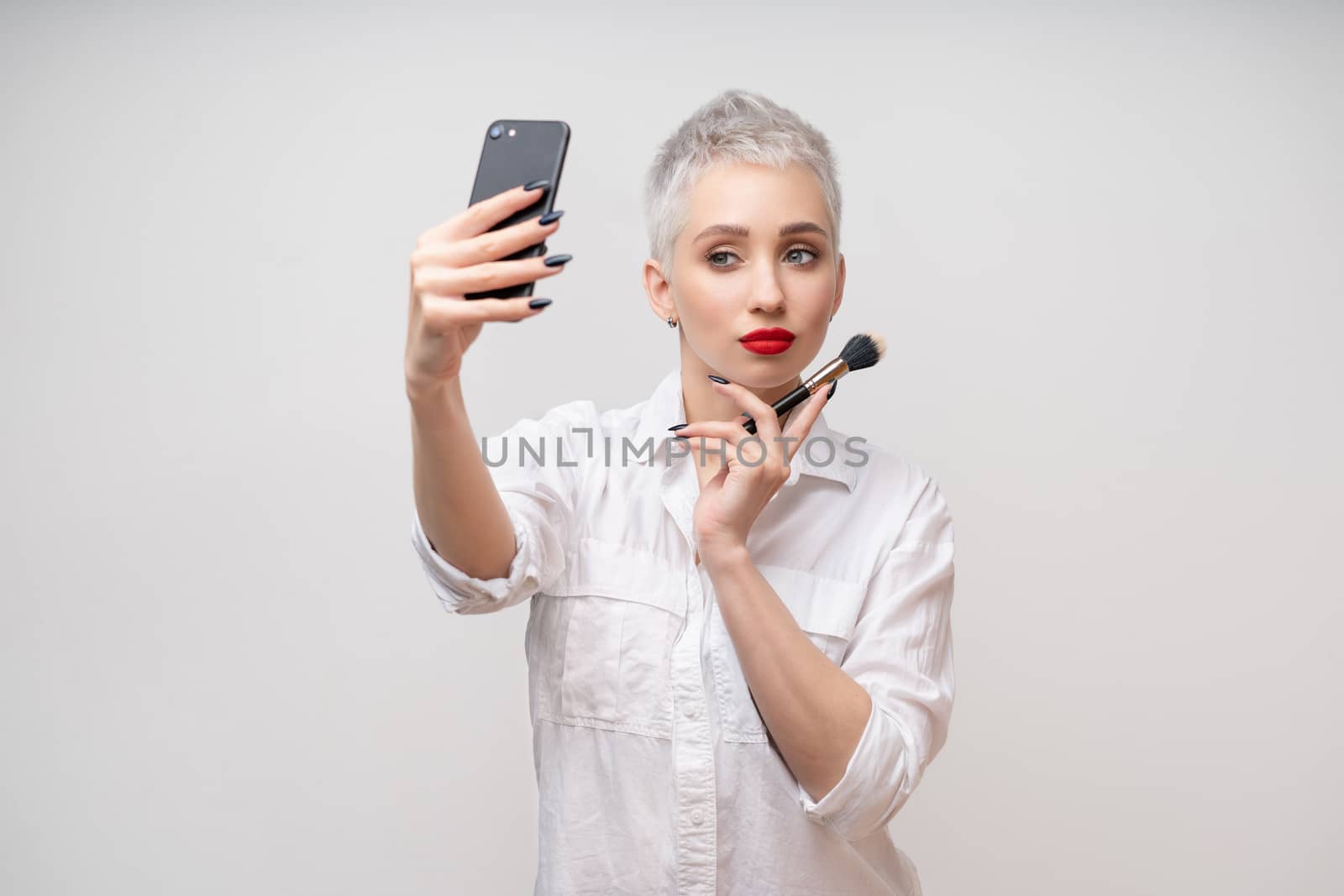Studio portrait of beautiful trendy girl with short hair and make up holding arm on waist and taking selfie via cell phone over white isolated background by andreonegin