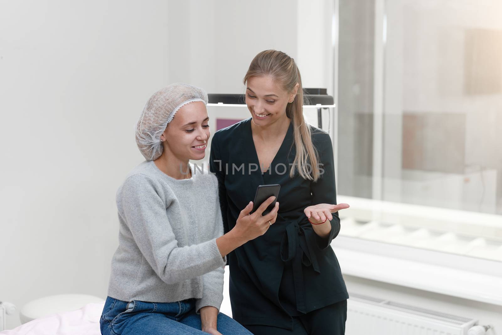 Cosmetology cabinet client sitting on couch. Beautician stands show smartphone display and smiles. Cosmetologist patients discuss choose treatment options. Preparation for procedure permanent makeup.