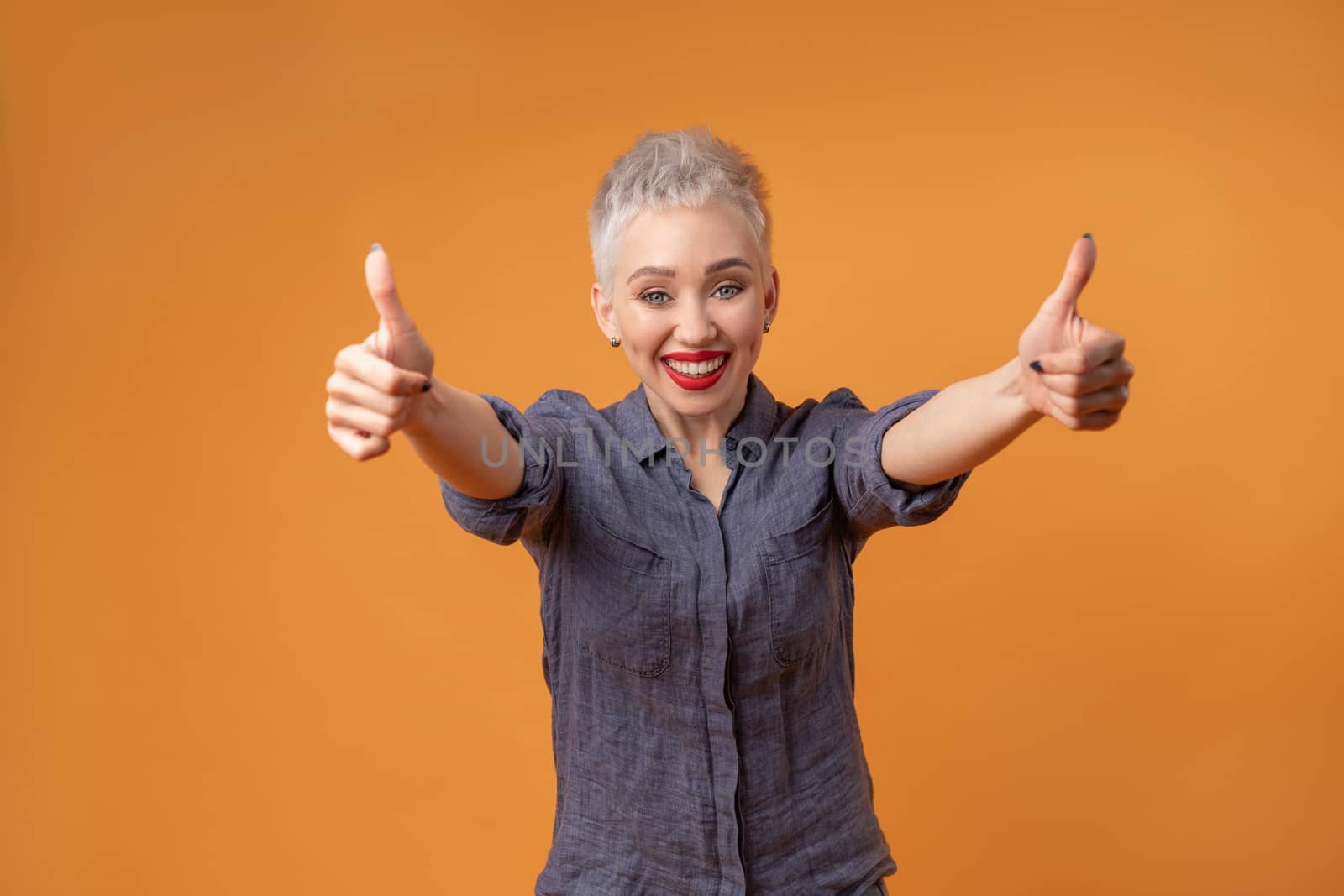 Portrait of young girl with blond short hairstyle looking at camera and laugh showing thumbs up on two hands on orange background with copy space. Woman surprised and smiling. positive emotion