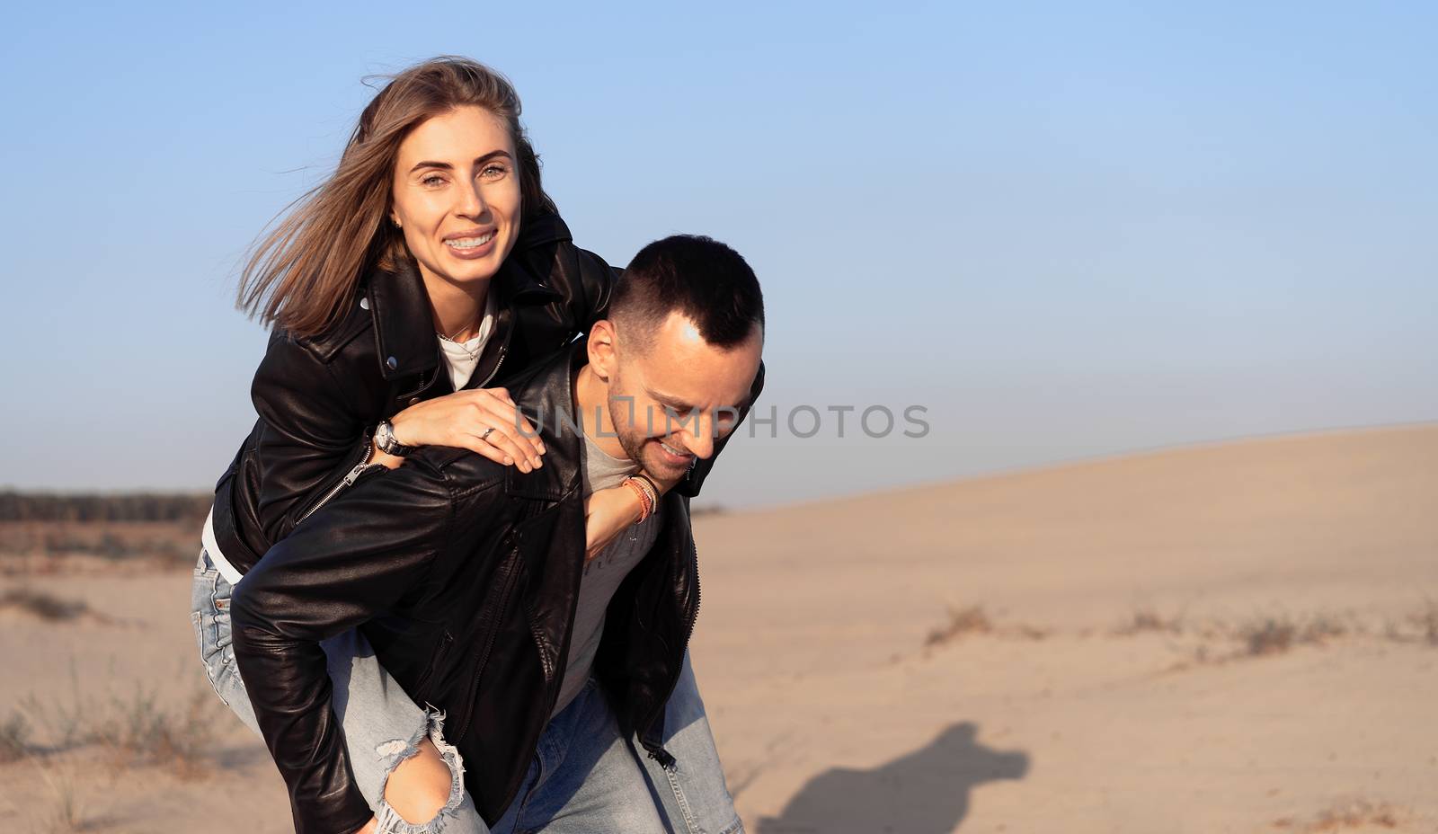 Happy and cute adorable adult couple leather jacket and jeans man with woman girlfriend on piggy back, have fun play, laugh,smile and jump on sunset at desert crazy in love, emotions and relationship