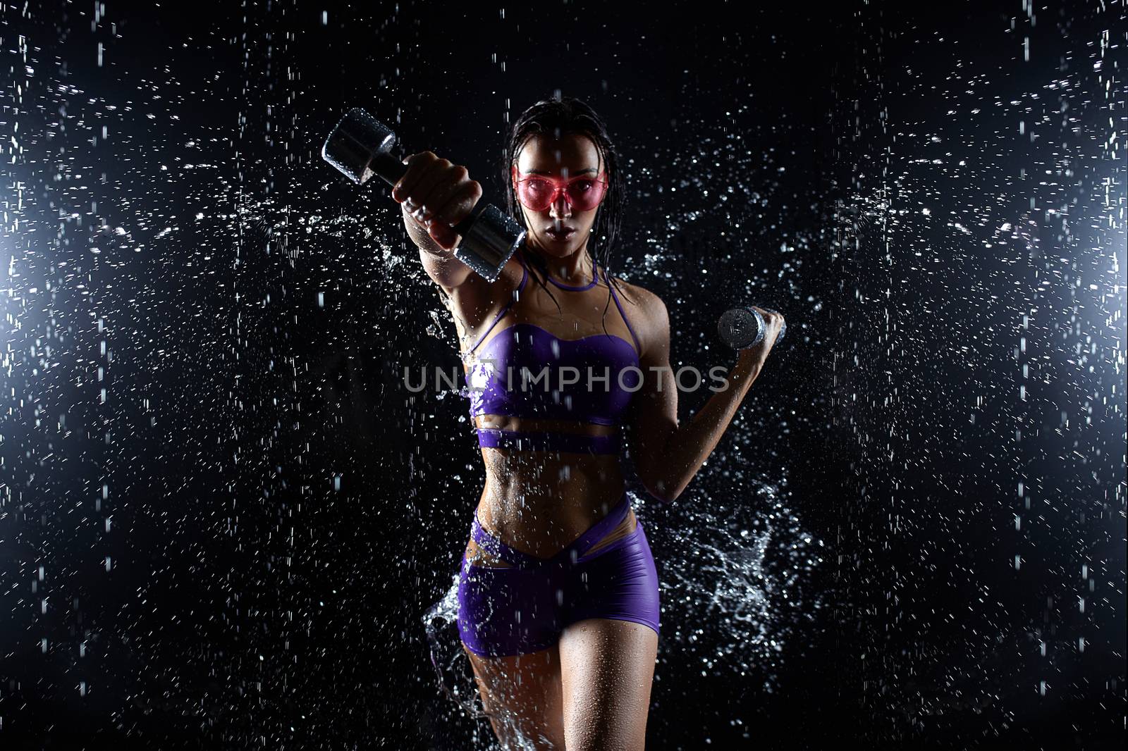 Beautiful young girl in purple sportswear poses with dumbbells in aqua studio. Drops of water spread about her fitness body. The perfect figure on the background of water splashes.
