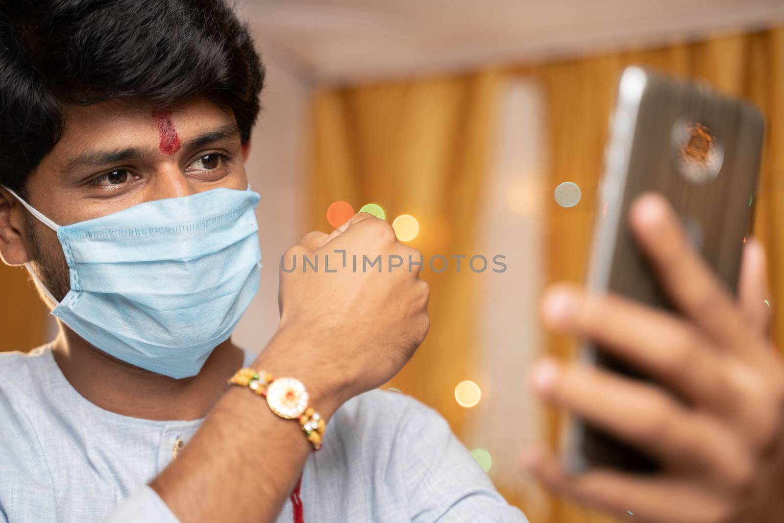 Man in medical mask busy on mobile phone and showing Rakhi or RakshaBandhan to sister or family friends at festival ceremony during coronavirus or covid-19 pandemic at home with decoration lights. by lakshmiprasad.maski@gmai.com