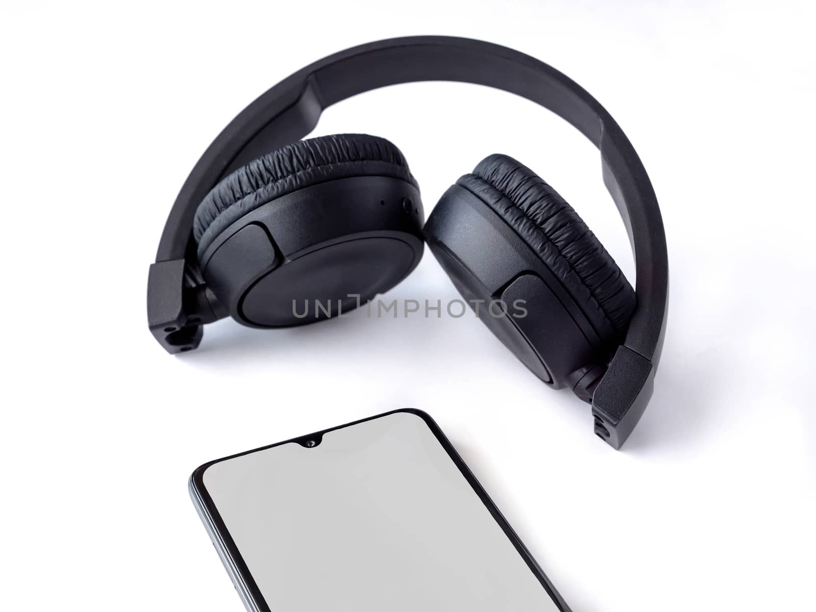 Black wireless headphone and mobile smartphone with a blank screen mockup lay on the surface of a white background. Top view closeup with selective focus and copy space. Music concept.