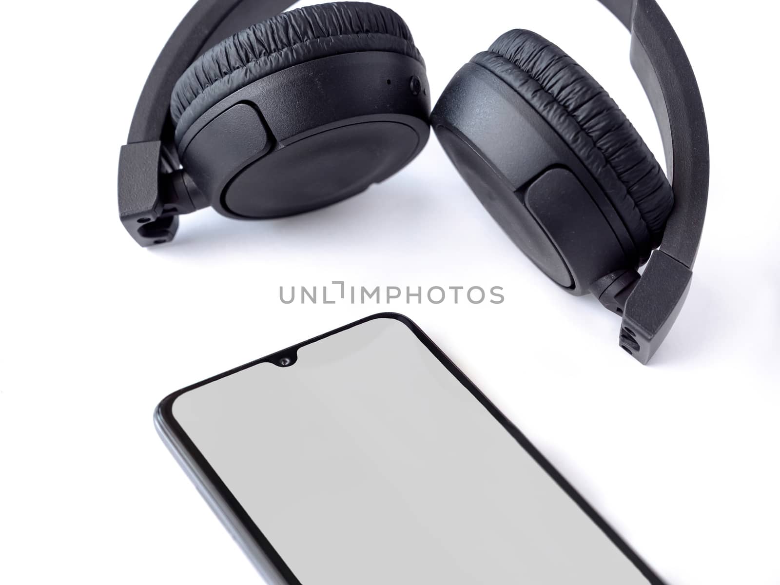Black wireless headphone and mobile smartphone with a blank screen mockup lay on the surface of a white background by wavemovies