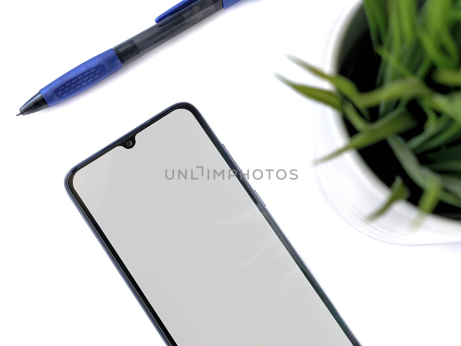 Modern minimalist workspace with black mobile smartphone mockup lies on the surface with a blank screen. Office desk table with a green plant and a pen. Top view flat lay closeup on white background.