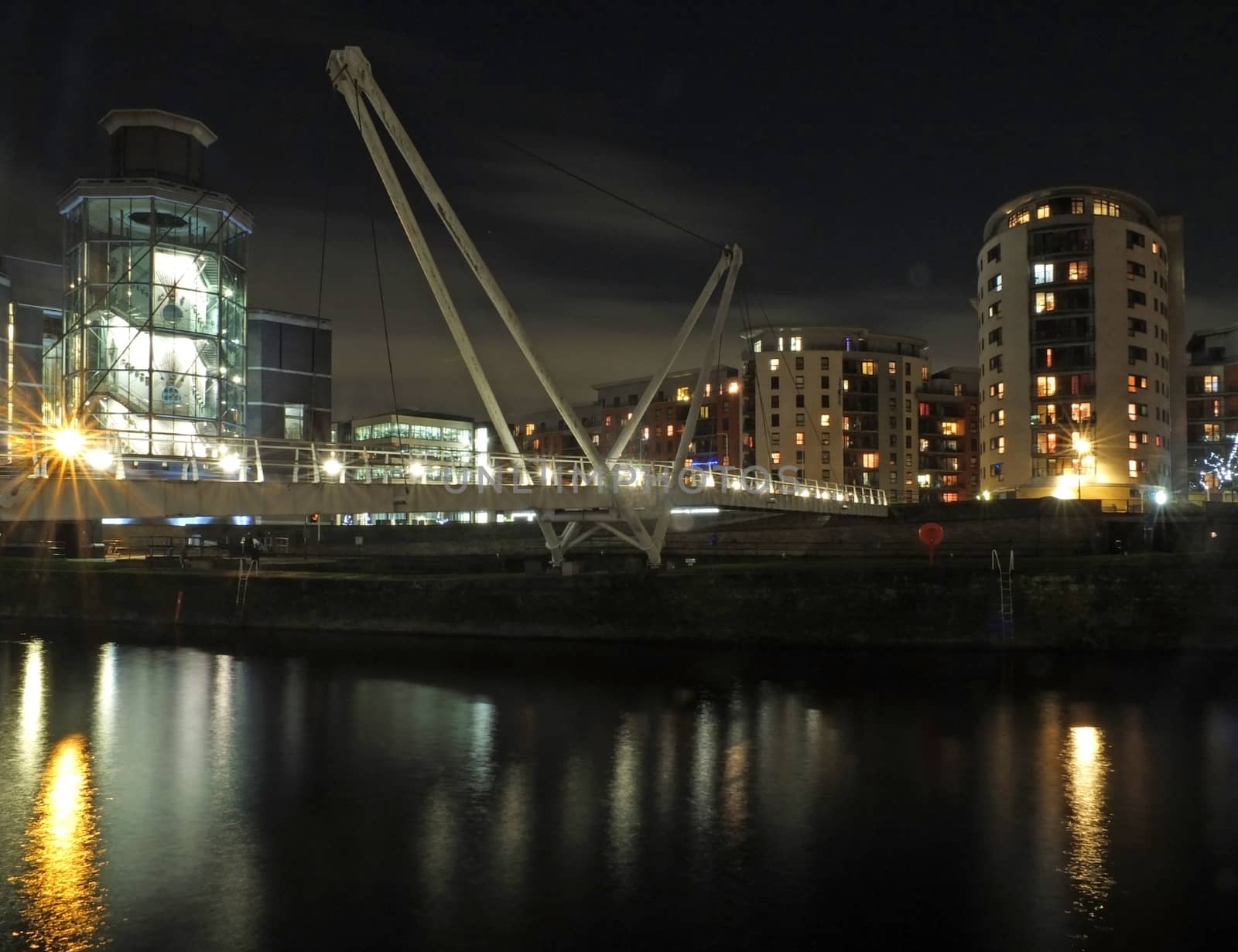 a cityscape view of the canal entrance to the clarence dock area of leeds with a pedestrian bridge crossing the water with reflections of lights and buildings against a night sky with clouds