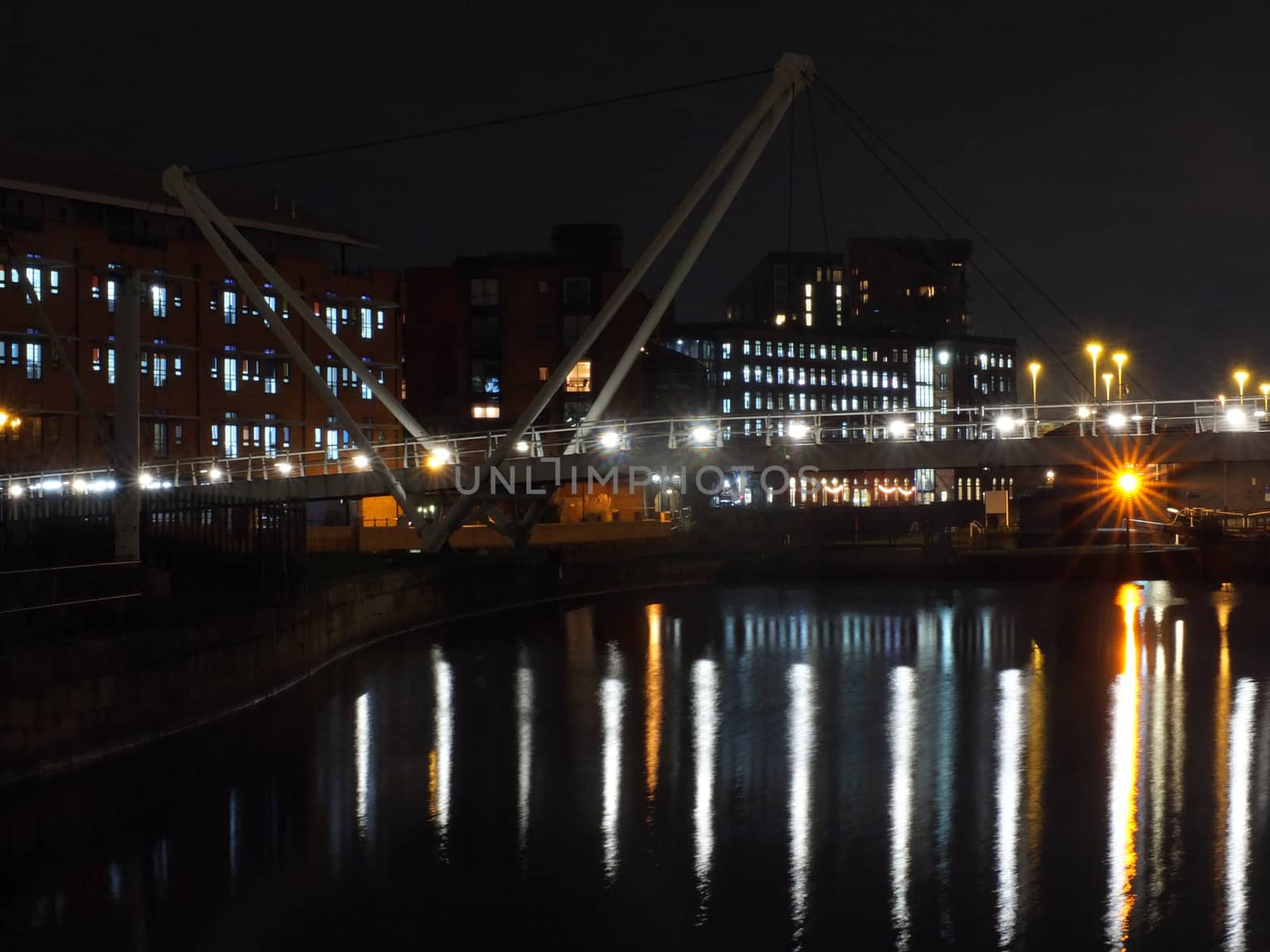 a cityscape view of the canal entrance to the clarence dock area of leeds with a pedestrian bridge crossing the water with reflections of lights and buildings against a night sky with clouds