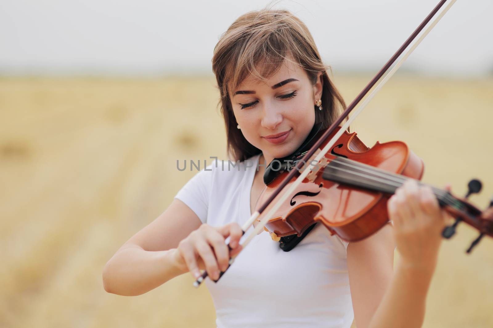 Beautiful romantic girl with loose hair playing the violin in the field after the harvest. Square sheaves of hay in the field. Violin training