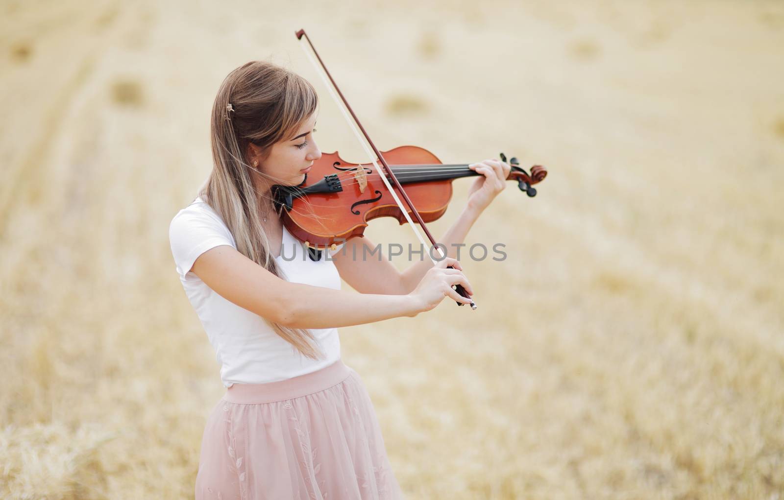 Beautiful romantic girl with loose hair playing the violin in the field after the harvest. Square sheaves of hay in the field. Violin training