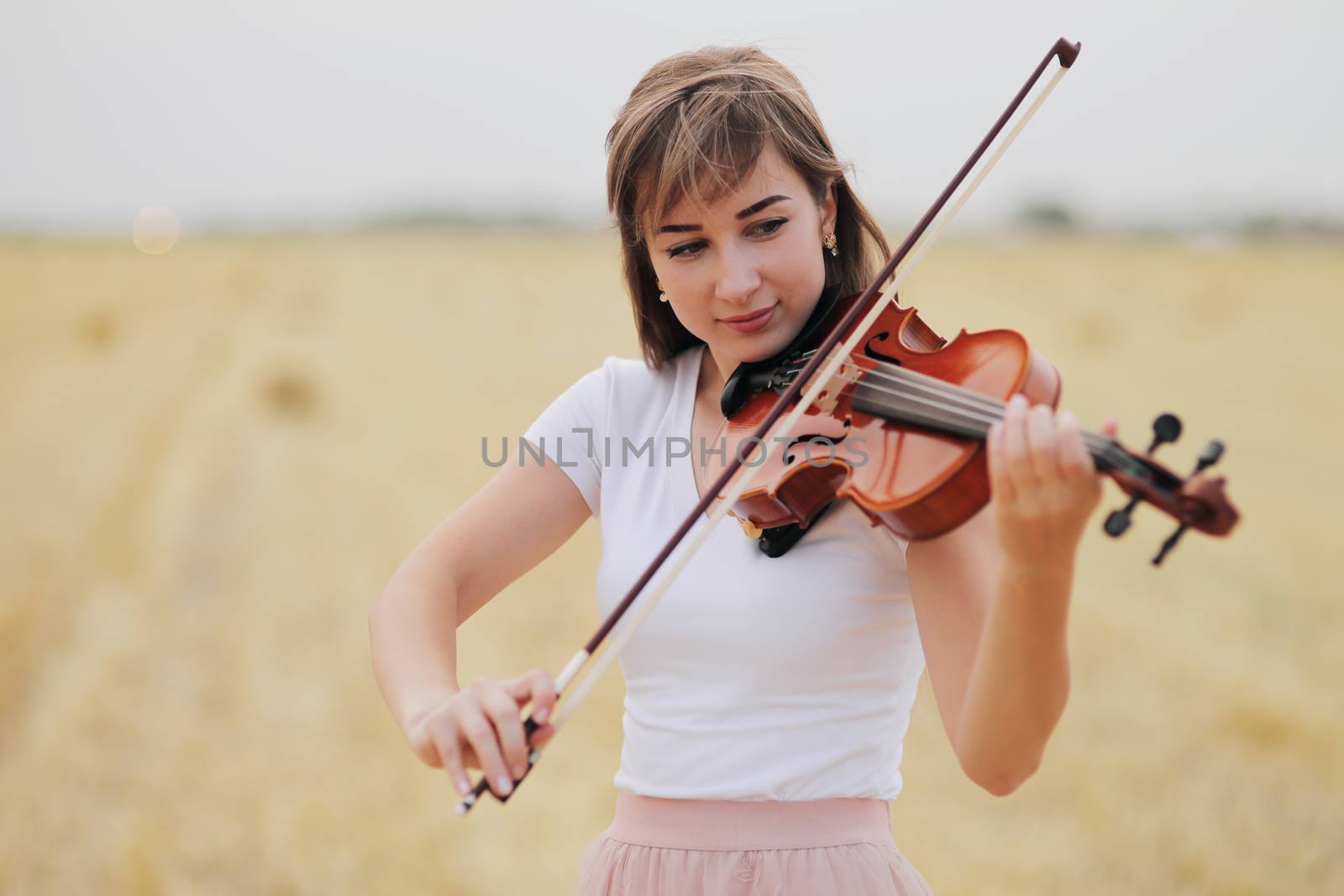Beautiful romantic girl with loose hair playing the violin in the field after the harvest. Square sheaves of hay in the field. Violin training