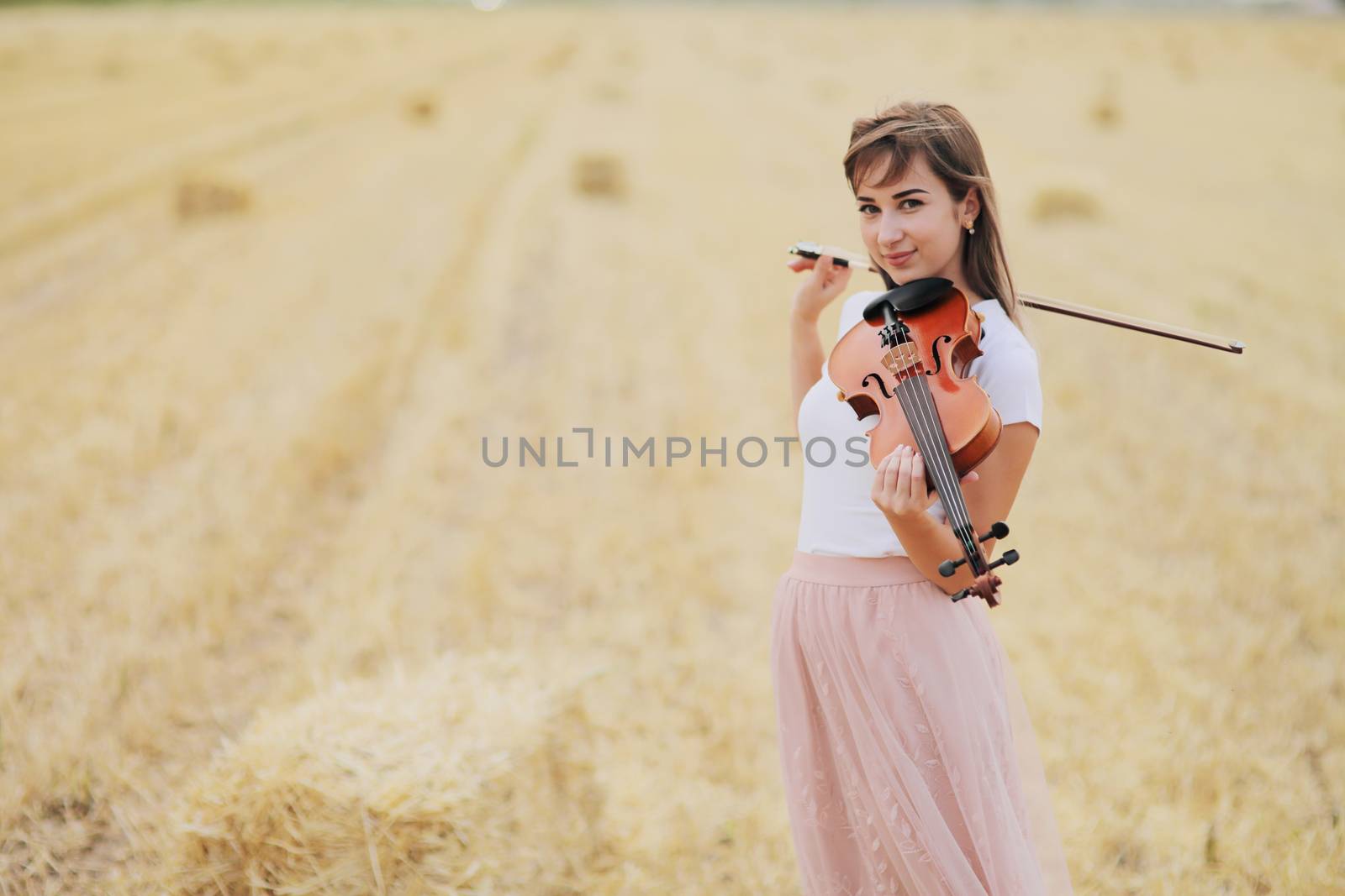 Beautiful romantic girl with loose hair playing the violin in the field after the harvest. Square sheaves of hay in the field. Violin training