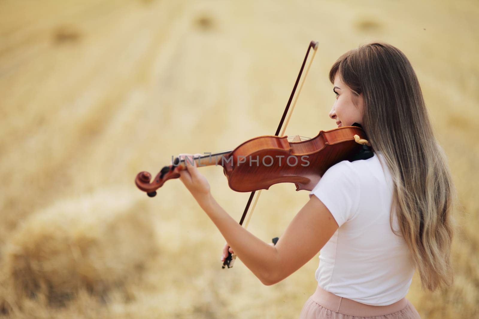 Beautiful romantic girl with loose hair playing the violin in the field after the harvest. Square sheaves of hay in the field. Violin training