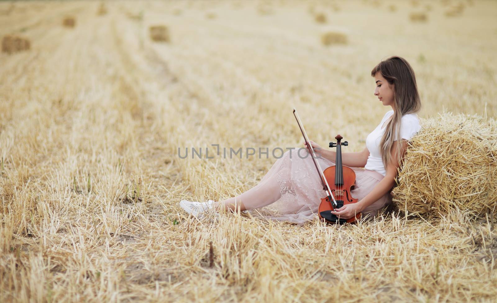 Romantic young woman with flowing hair, holding a violin in her hand in a field after harvest. Square sheaves of hay in the field. Violin training
