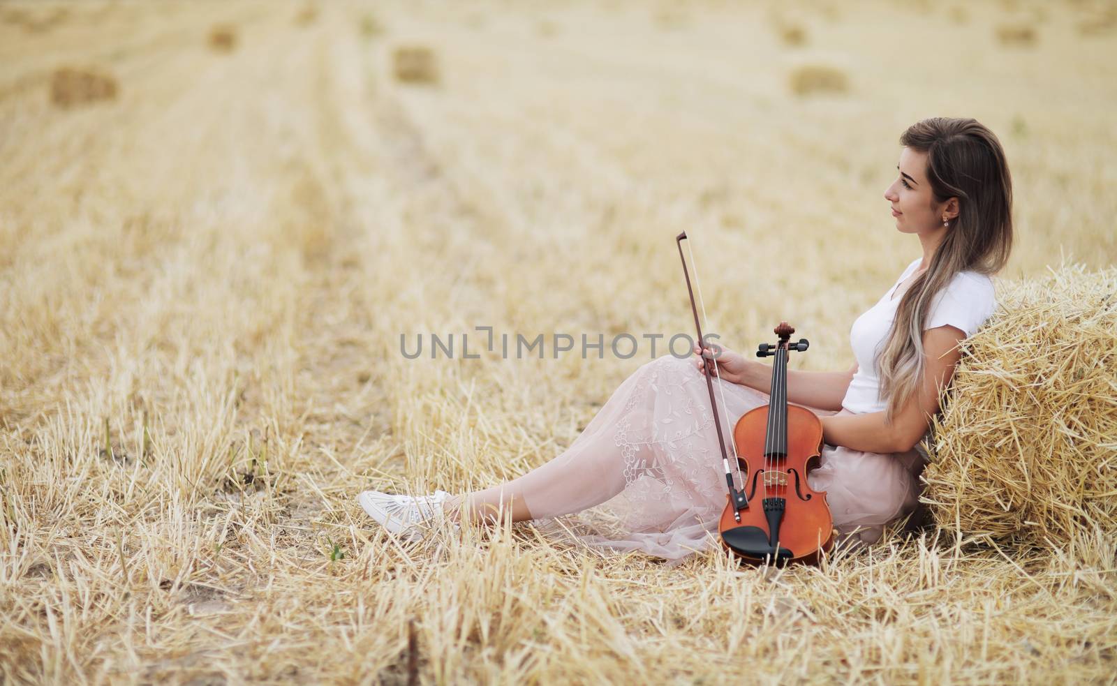 Romantic young woman with flowing hair, holding a violin in her hand in a field after harvest. Square sheaves of hay in the field. Violin training