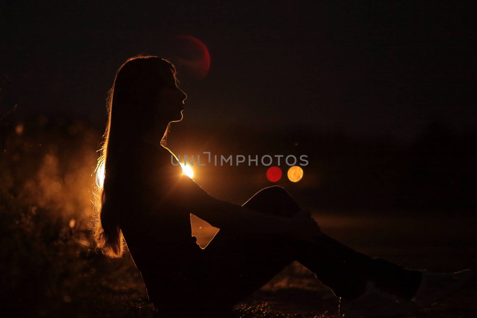 Silhouette of young slender woman in the backlight of car headlights on the auto road at night. The girl is hitchhiking.