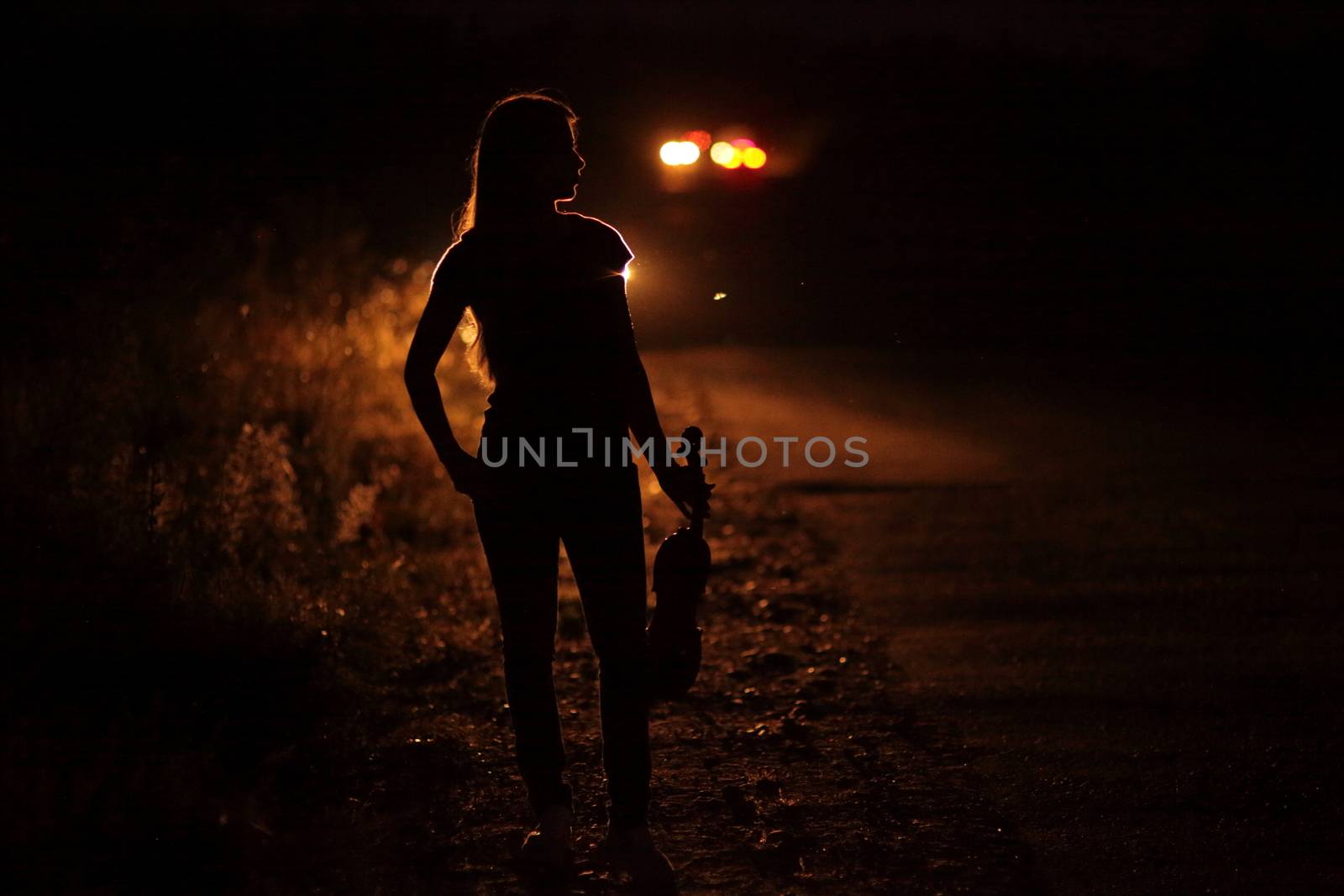 Silhouette of a slender young woman with violin in car headlights illumination. A young slender girl in the backlight of car headlights on the auto road at night. The girl is hitchhiking.