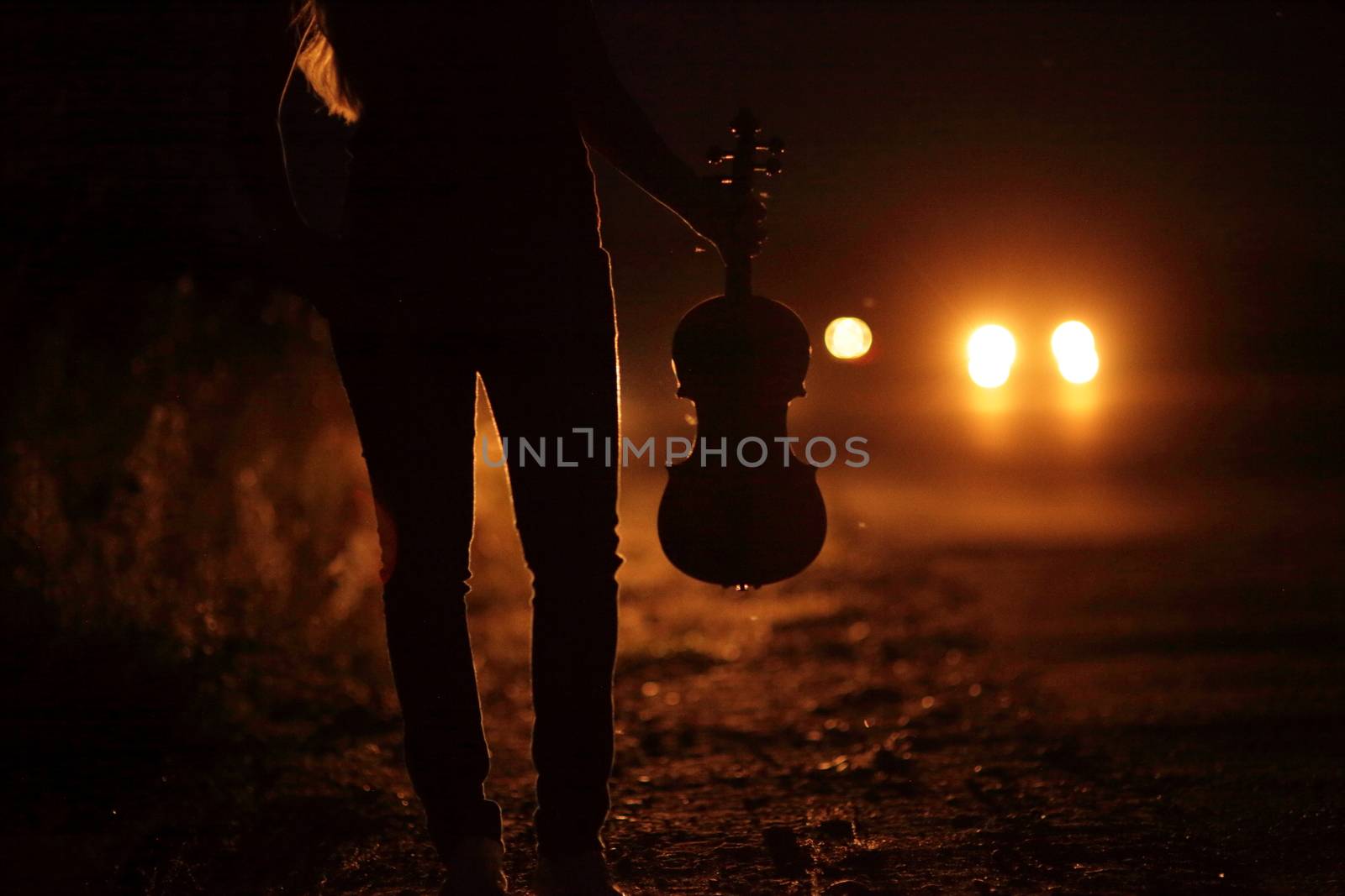 Silhouette of a slender girl's legs and a violin in the backlight of headlights. A young slender woman in the backlight of car headlights on the auto road at night. The girl is hitchhiking.