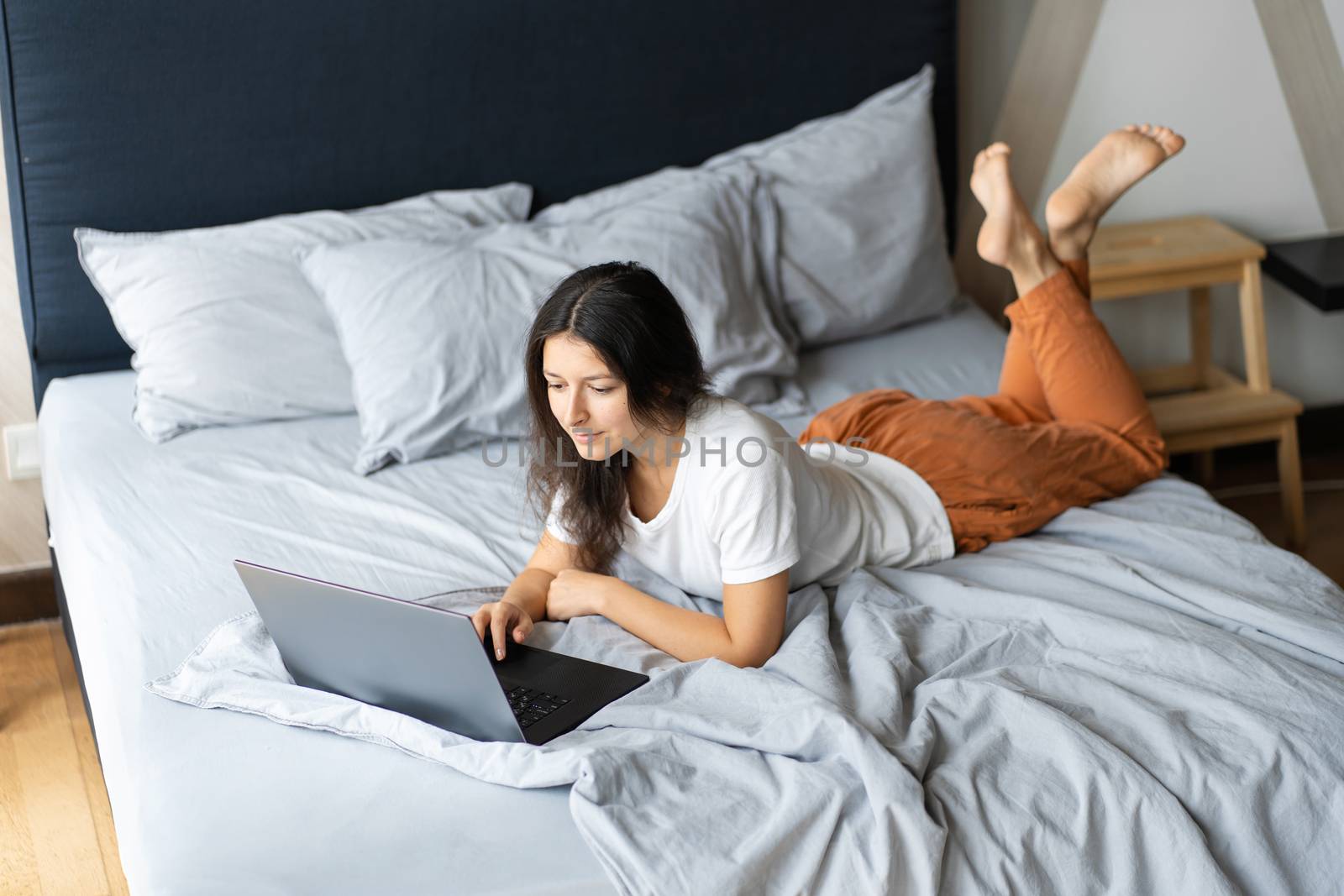 Beautiful young brunette girl with a laptop lying on the bed. Stylish modern interior. A cozy workplace. Shopping on the Internet. by Try_my_best