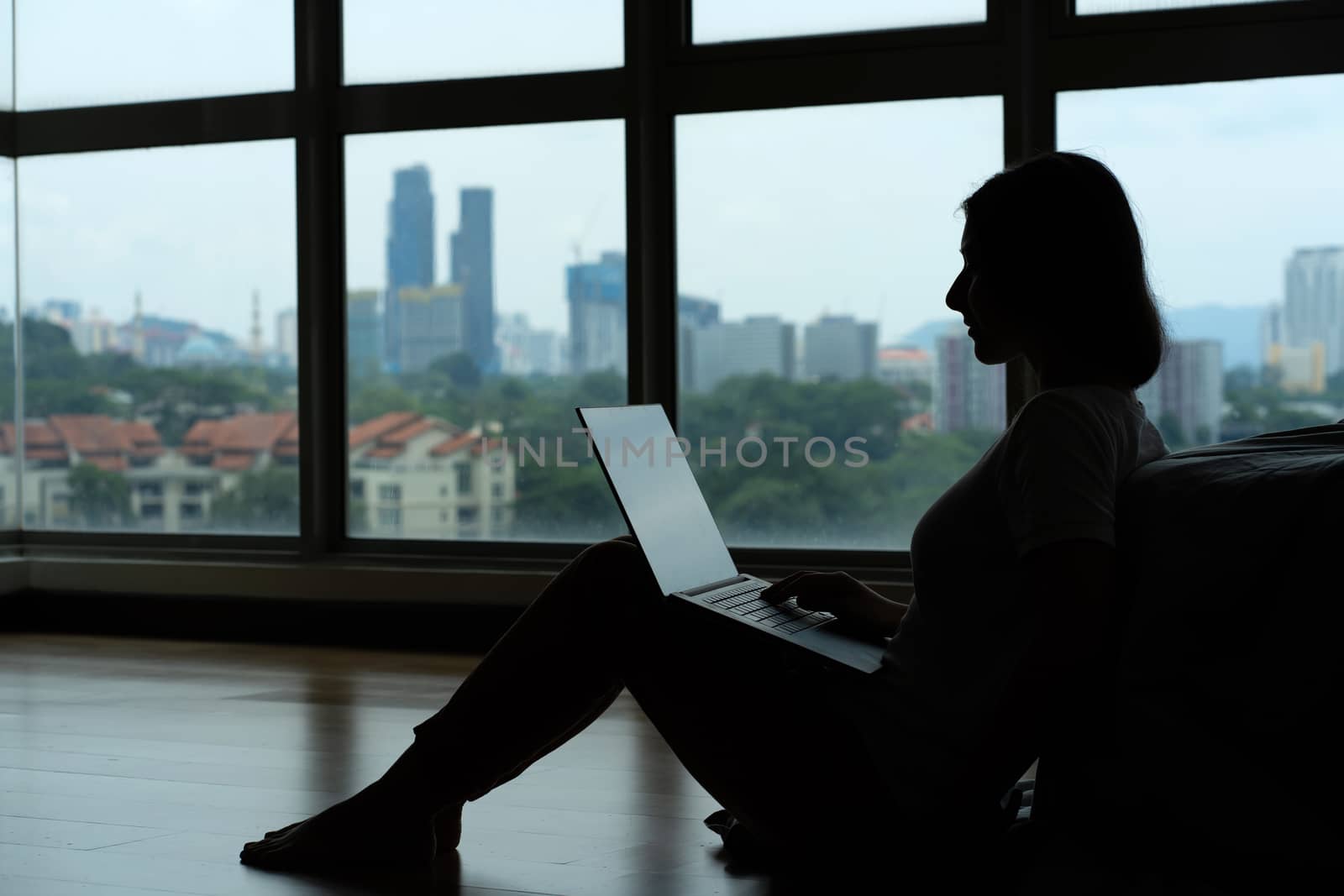 Siluet girl working on a laptop and drinking coffee, sitting on the floor near the bed by the panoramic window with a beautiful view from the high floor. Stylish modern interior.