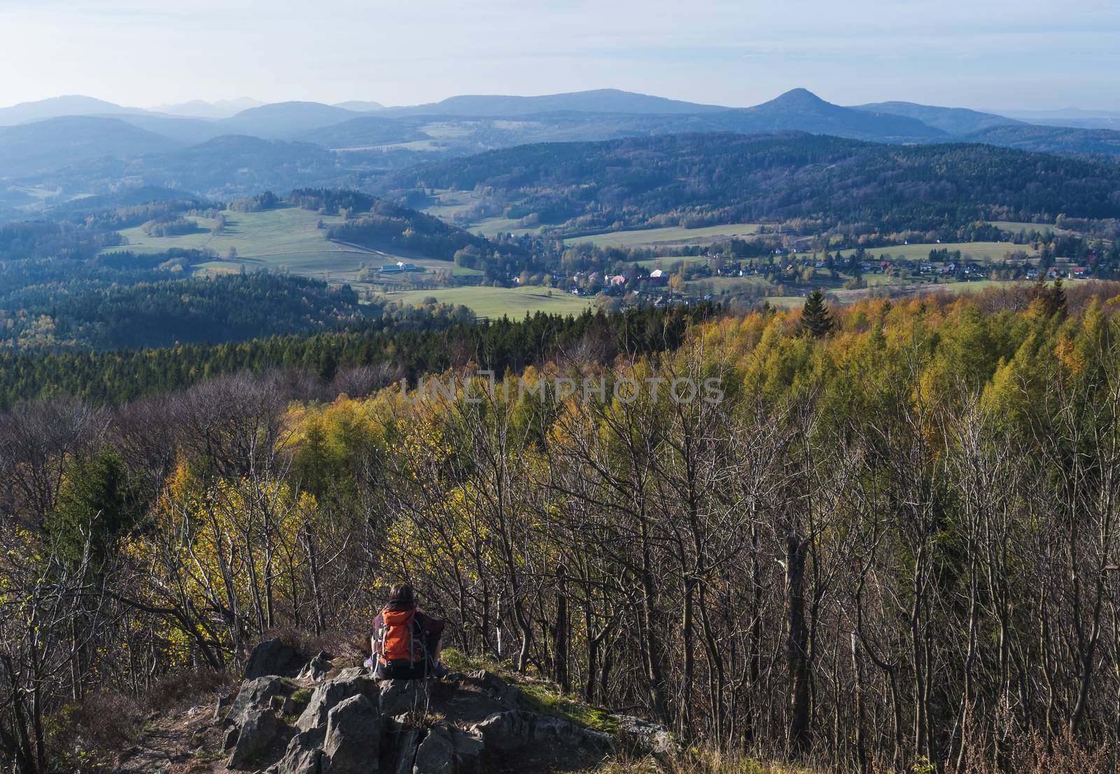 Young woman hiker takeing pictutre of Luzicke hory panorama from Hochwald Hvozd view-point of the Lusatian Mountains with autumn colored deciduous and coniferous tree forest and green hills, golden hour light.