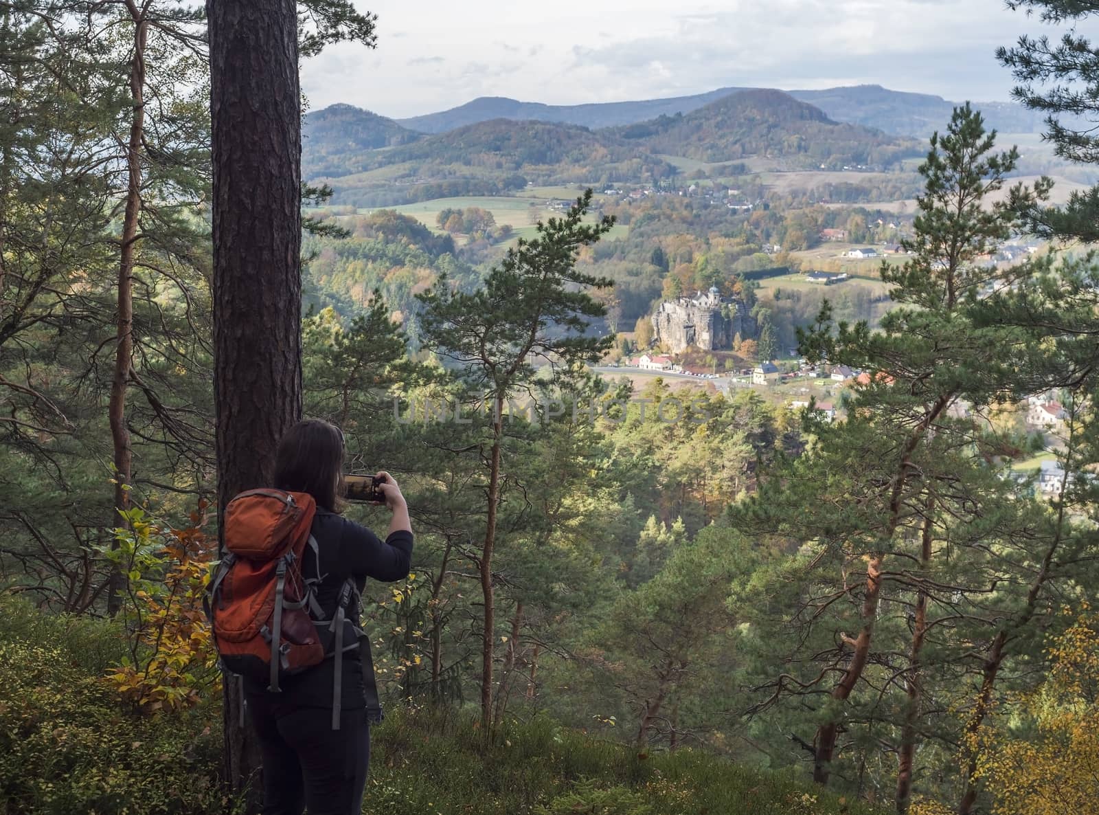 Young woman takeing cell phone picture of view on village Sloup v cechach with mediaval sand stone castle in luzicke hory, Lusatian Mountains with autumn colored tree forest and green hills, blue sky, white clouds.