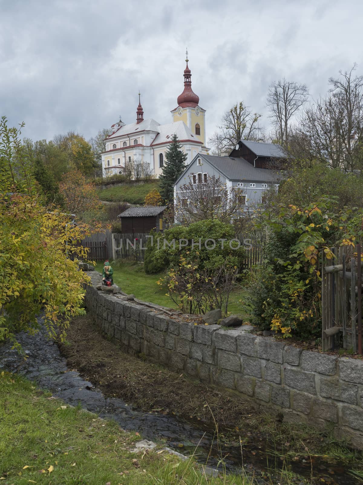 view on church and houses in village Sloup v cechach in luzicke hory, Lusatian Mountains with autumn colored trees and small water stream, blue sky, white clouds.