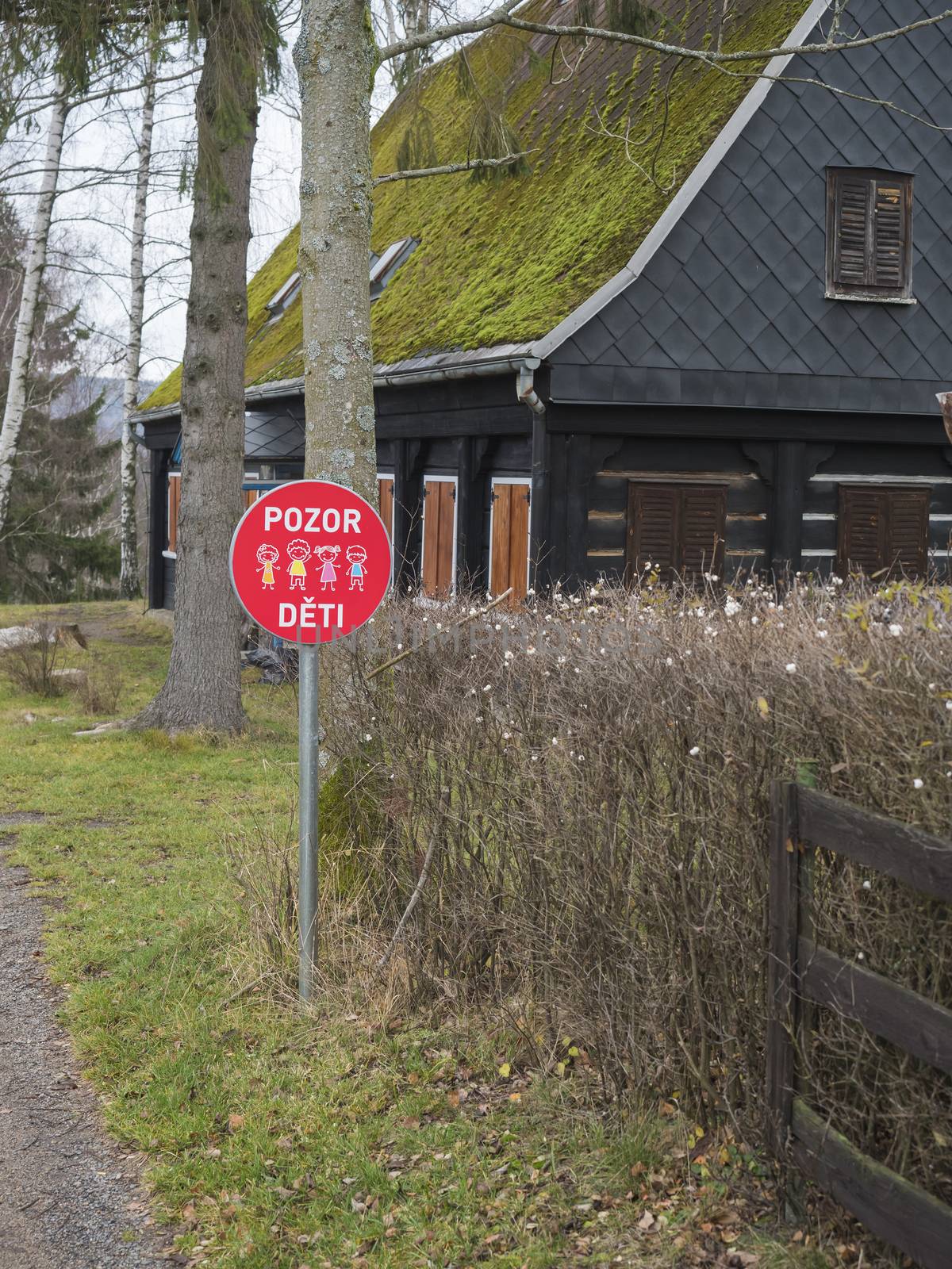 Red traffic sign with czech text: Pozor deti - Attention children in front of wooden timbered house cotttage with moss roof. by Henkeova