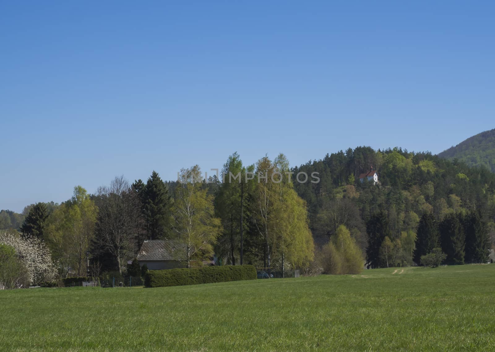 Spring landscape with view on village Marenice in Lusitian mountains with small chapel on hill, lush green grass meadow, deciduous and spruce tree forest and hills, blue sky background.
