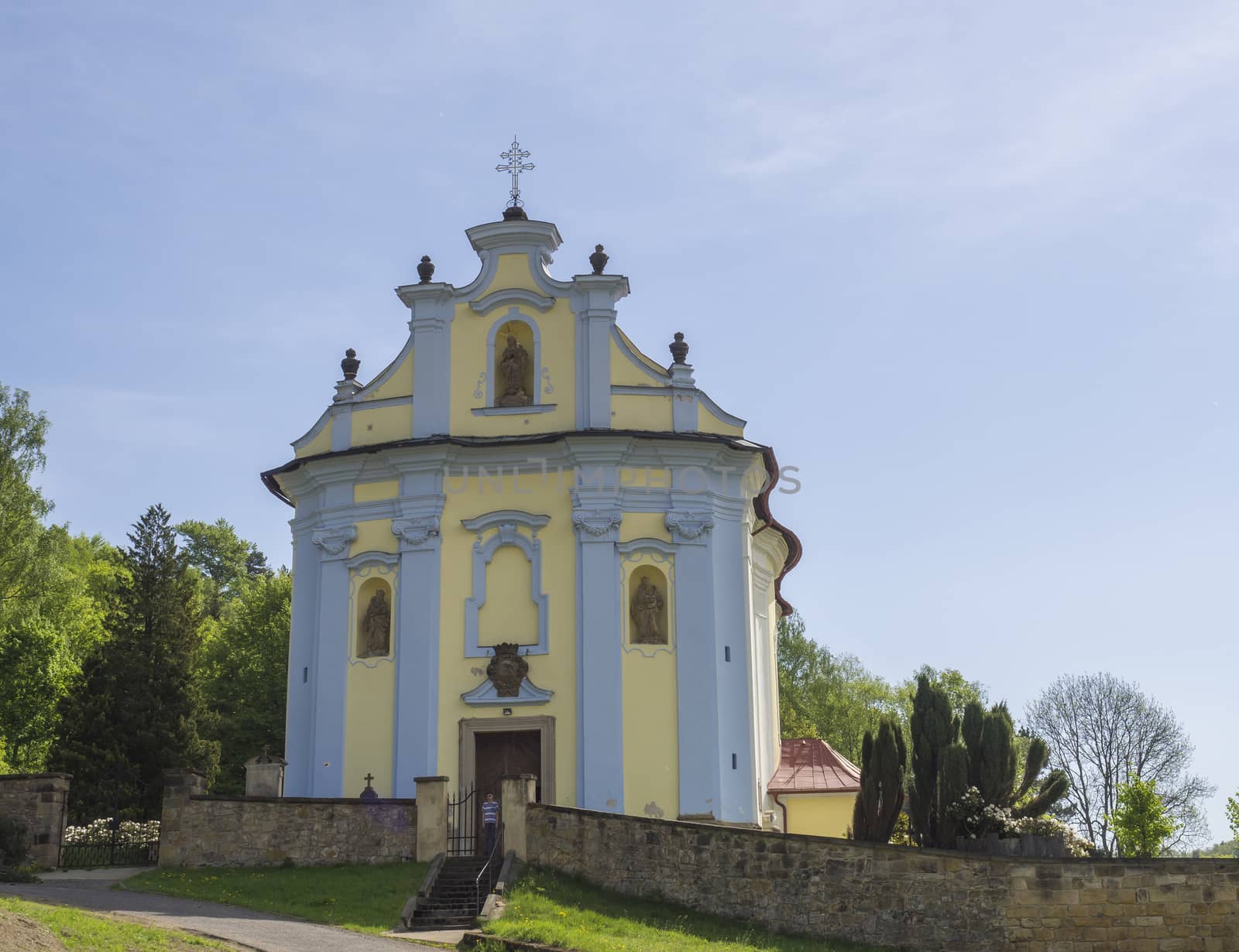 blue and yellow church in baroque style in horni prysk in czech republic with surrounding cemetery wall, boy standing at the door, blue sky background