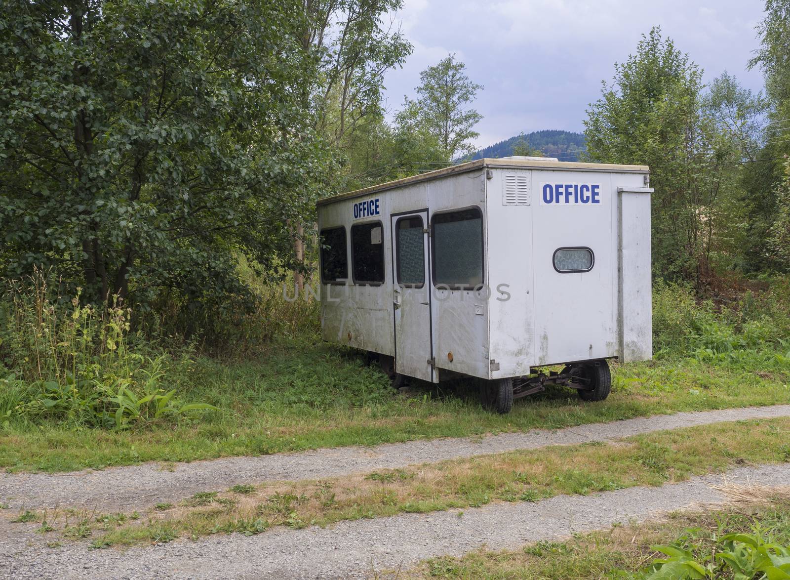 old small white trailer house with blue sign office, abandoned caravan in rural landscape near footpath, green grass and trees, blue sky