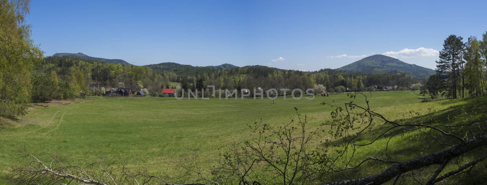 Panoramic landscape with view on village Marenice in Lusitian mountains with traditional wooden cottage and lush green grass meadow, deciduous and spruce tree forest and hills, spring, blue sky background.