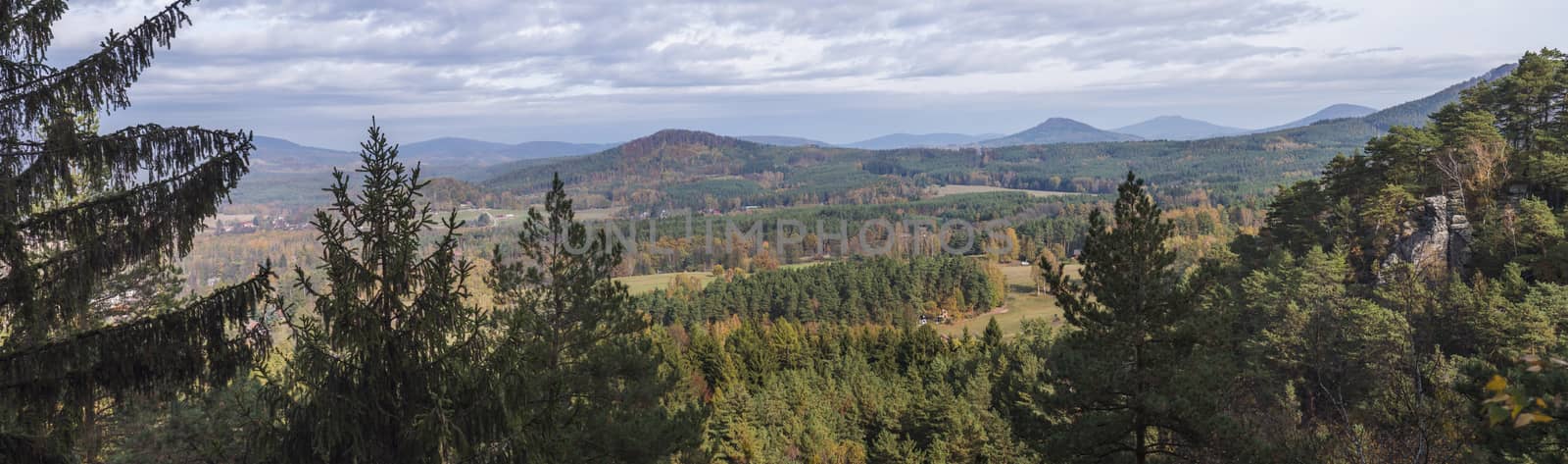 Panoramic view from Hrabencina vyhlidka on village Sloup v cechach in luzicke hory, Lusatian Mountains with autumn colored deciduous and coniferous tree forest and green hills, blue sky, white clouds.