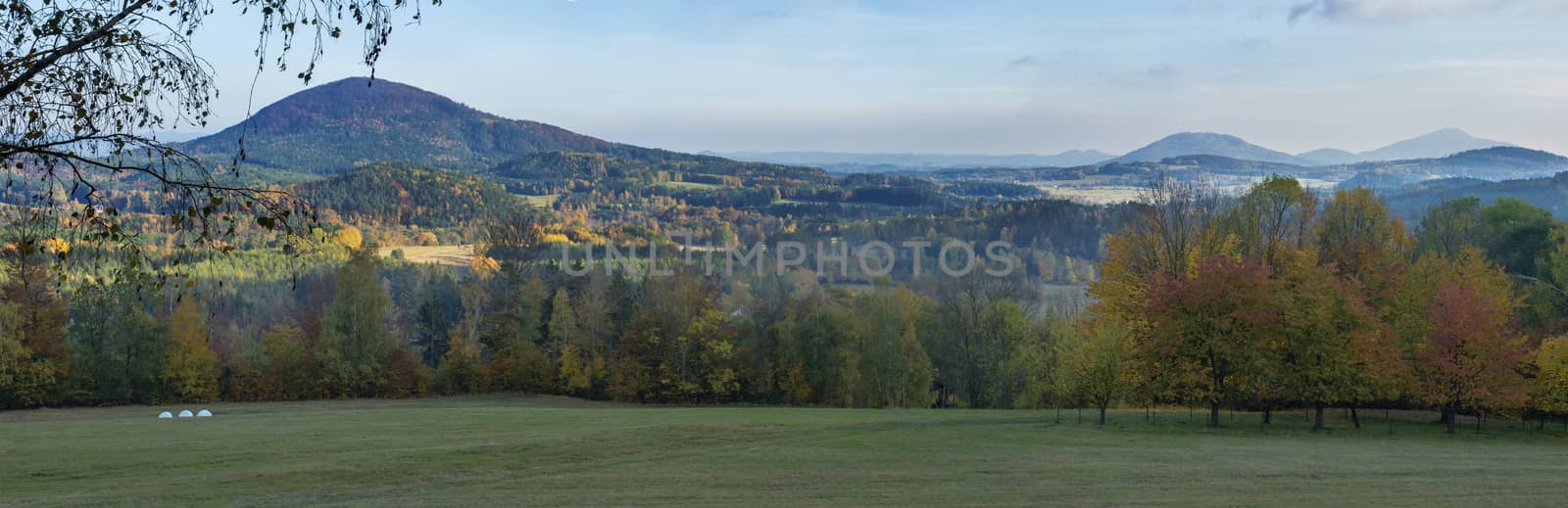 Panoramic Landscape in luzicke hory, Lusatian Mountains. Meadow, autumn colored deciduous and coniferous tree forest and green hills, blue sky background, golden hour.