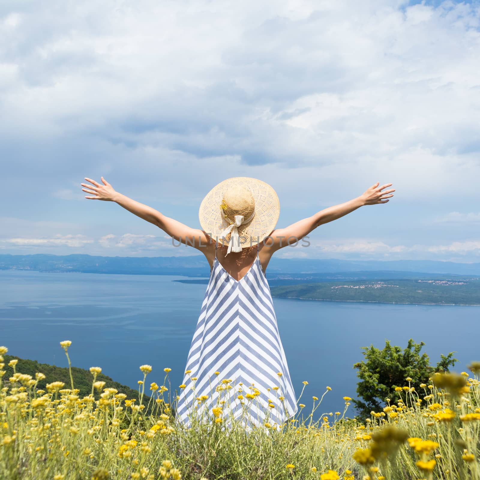 Rear view of young woman wearing striped summer dress and straw hat standing in super bloom of wildflowers, relaxing with hands up to the sky, enjoing beautiful view of Adriatic sea nature, Croatia by kasto