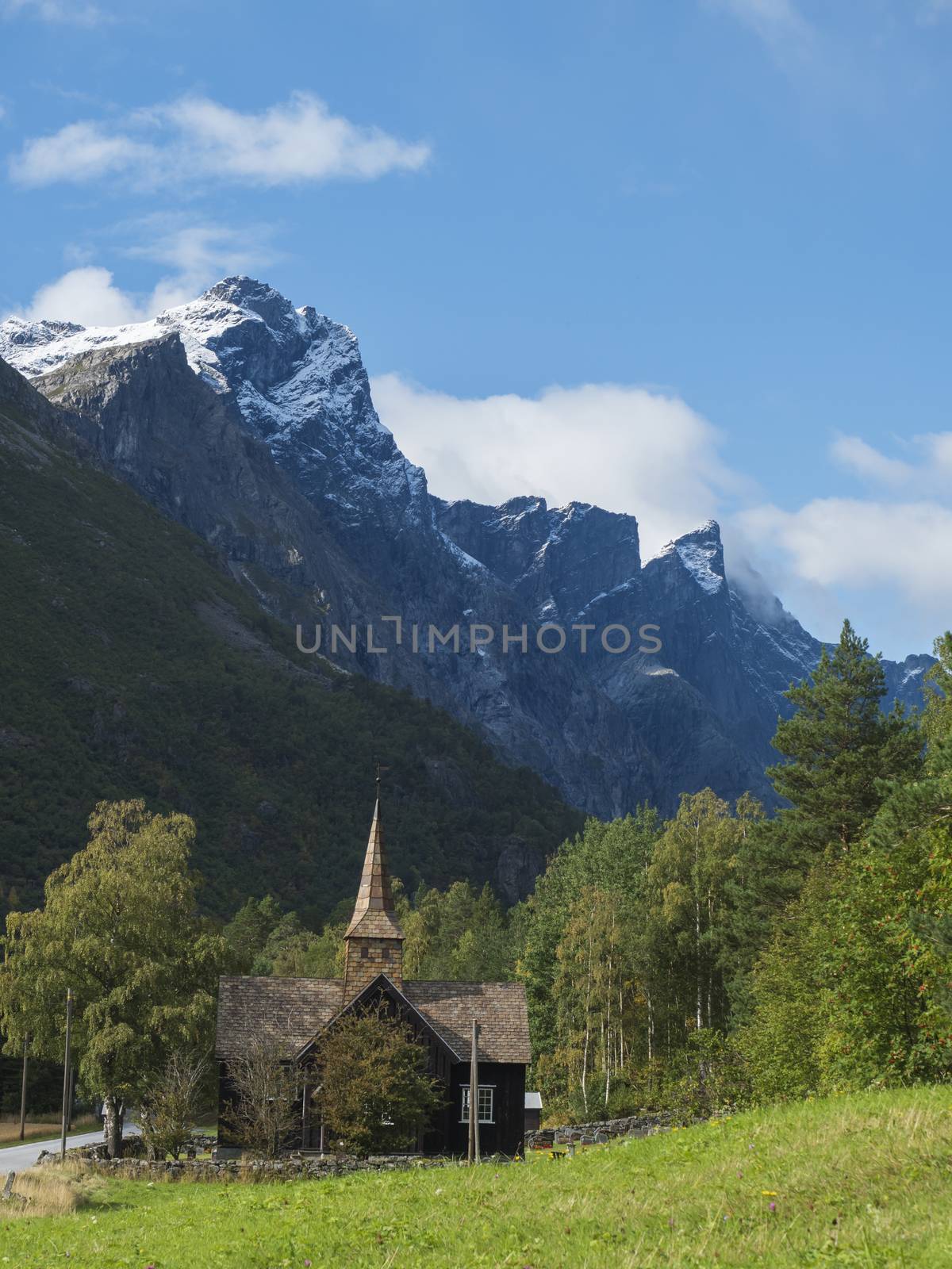 Kors Church, old wooden parish church in Rauma in Romsdal valley, Norway with road E136, green forest and mountain massif Trolltindene, Troll wall Trollveggen. Summer blue sky white clouds