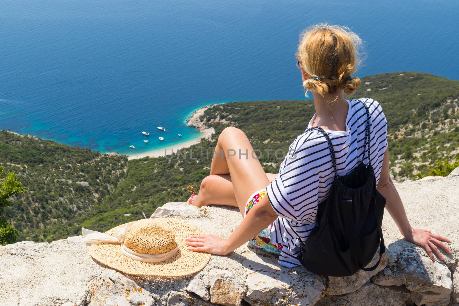 Active sporty woman on summer vacations sitting on old stone wall at Lubenice village, wearing straw hat and beach backpack enjoying beautiful coastal view of Cres island, Croatia.