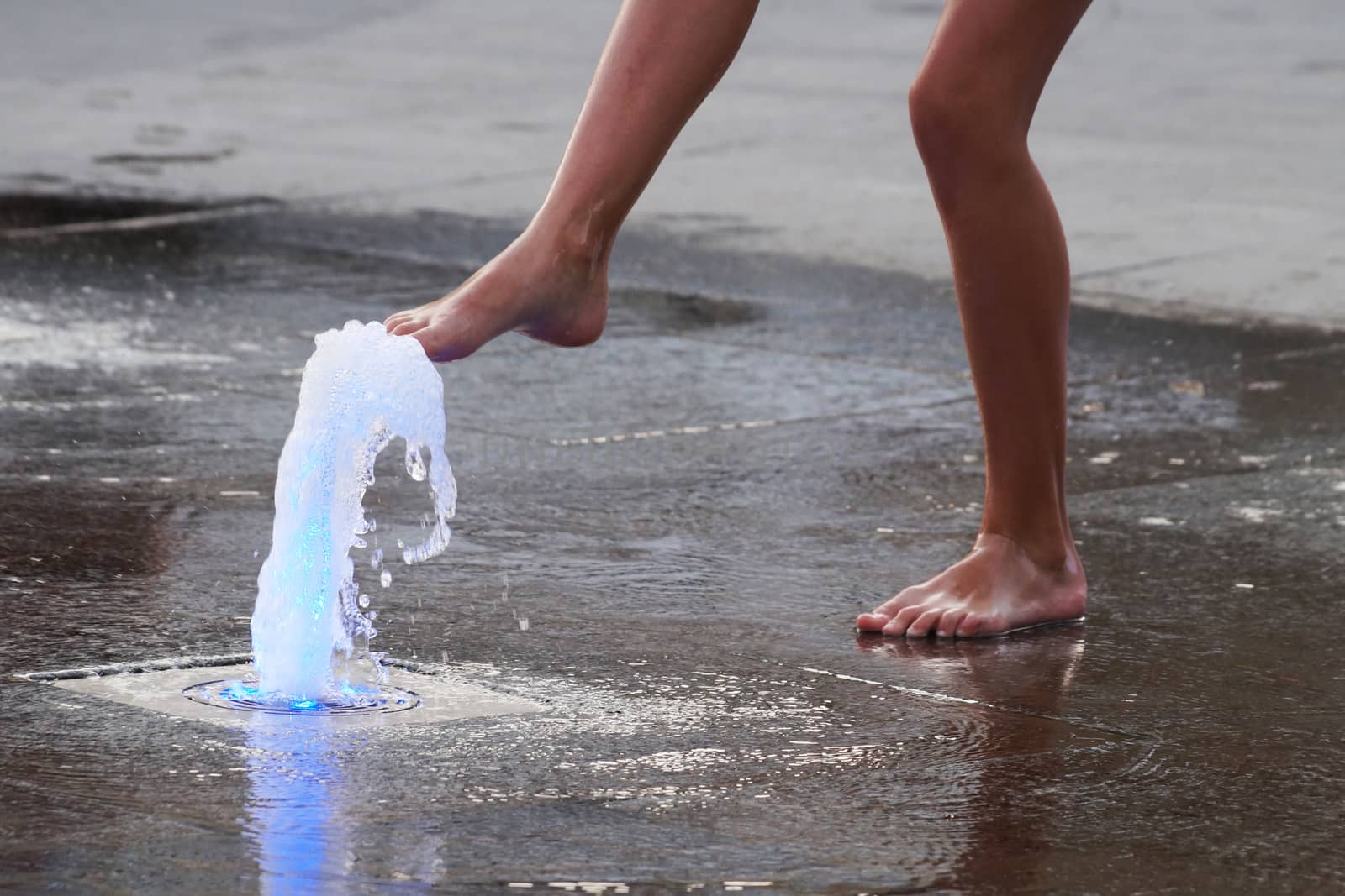 a girl touches a fountain on the sidewalk with her bare foot.