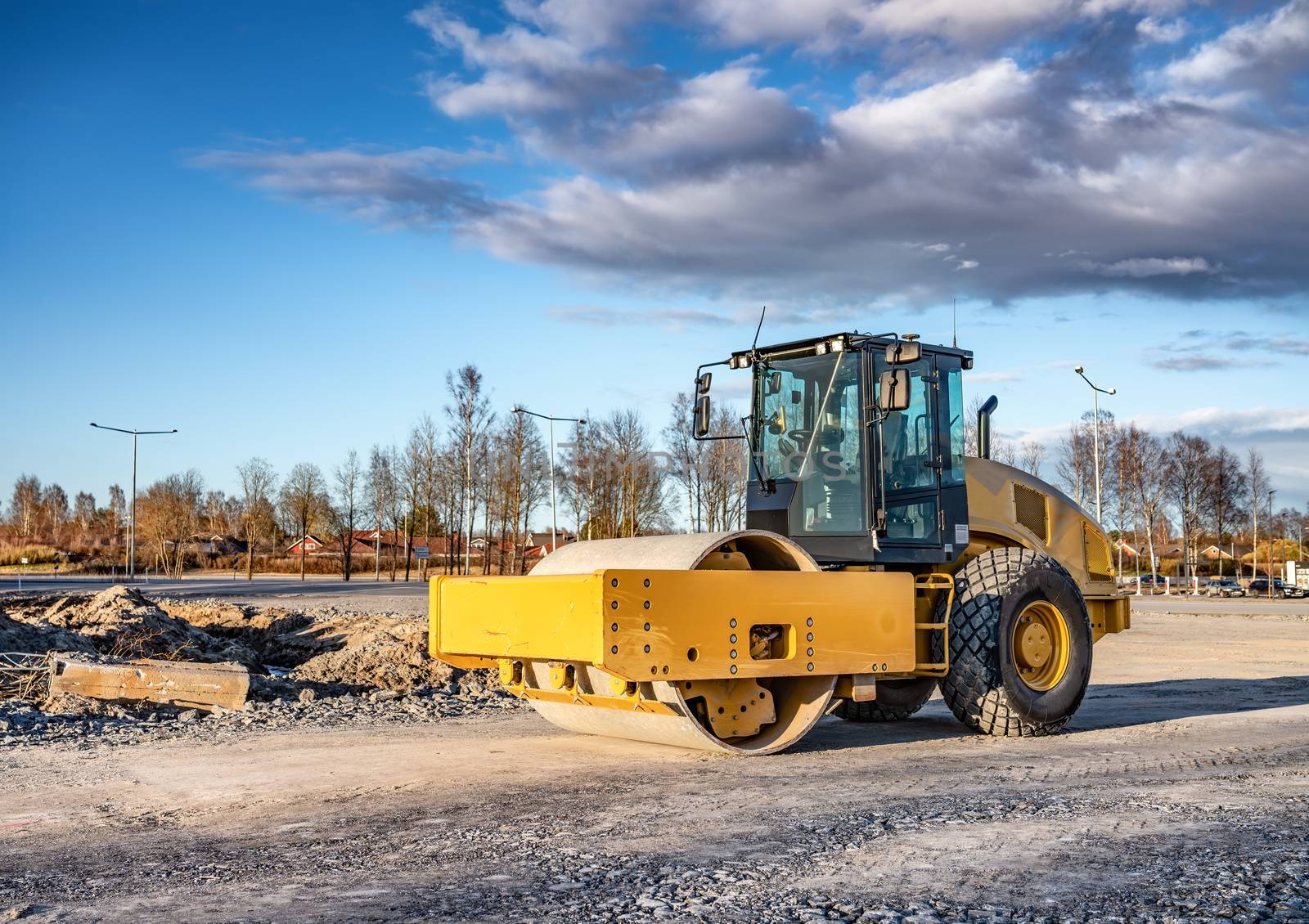 Eye catching yellow road roller with enclosed climate controlled cabin stands on not ready new road, stones, blue sky, clouds, front right side view. Clean shiny old heavy tractor by skydreamliner