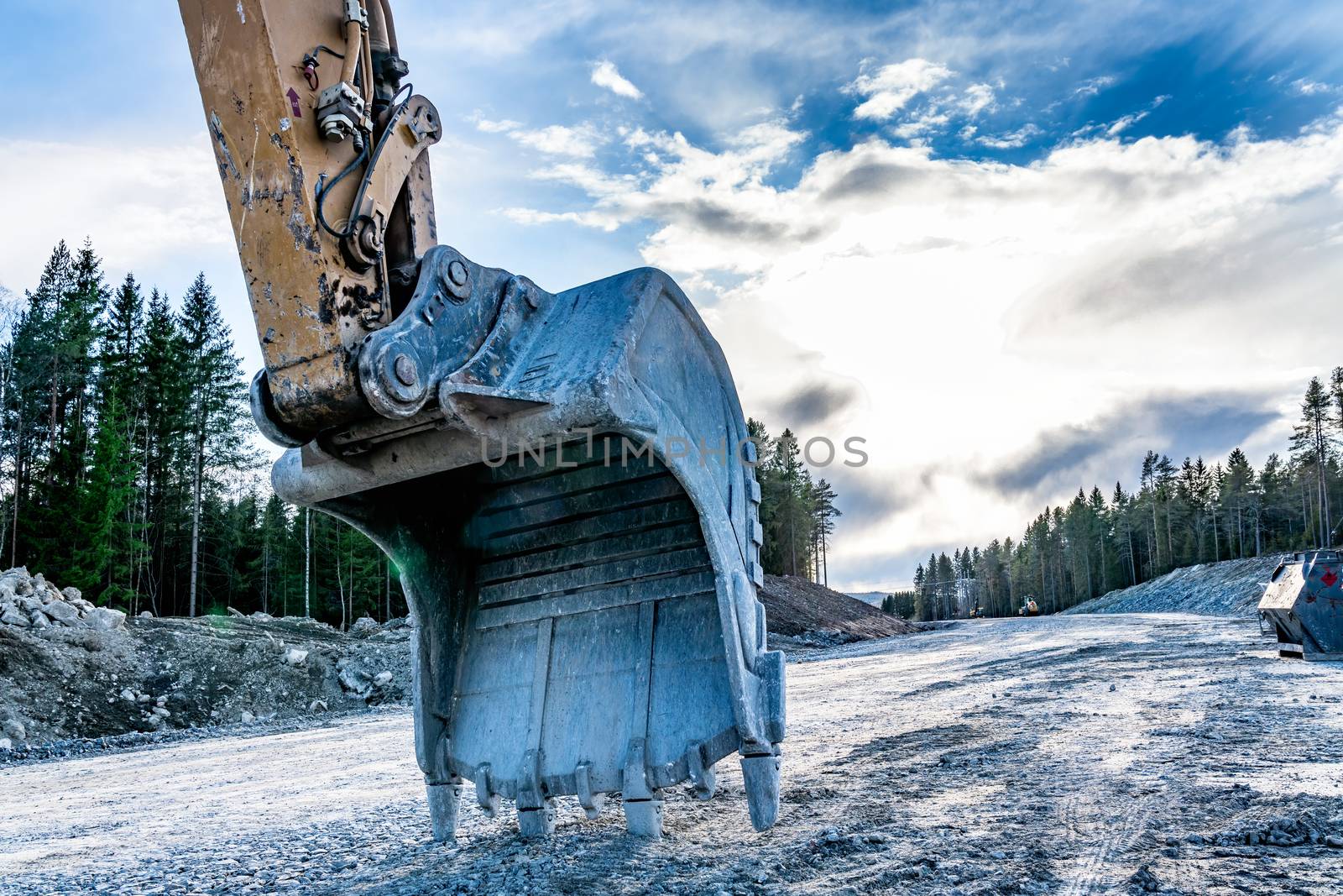 Close up back view at beautiful large excavator's scoop on new not yet ready road. Sunny evening with dark blue clouds, spring time, mixed forest with pile of big stones, dirt, sand at background.