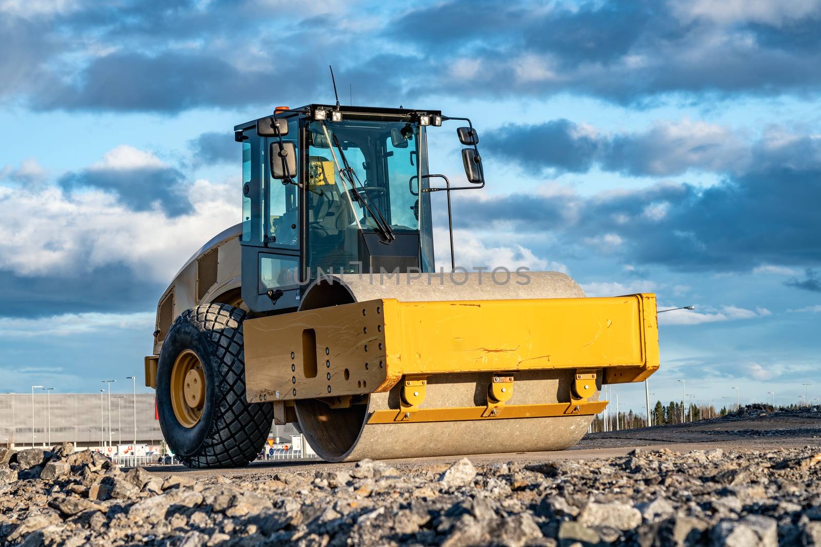 Eye catching yellow road roller with enclosed climate controlled cabin stands on not ready new road, stones, blue sky, clouds, front left side view. Clean shiny old heavy tractor