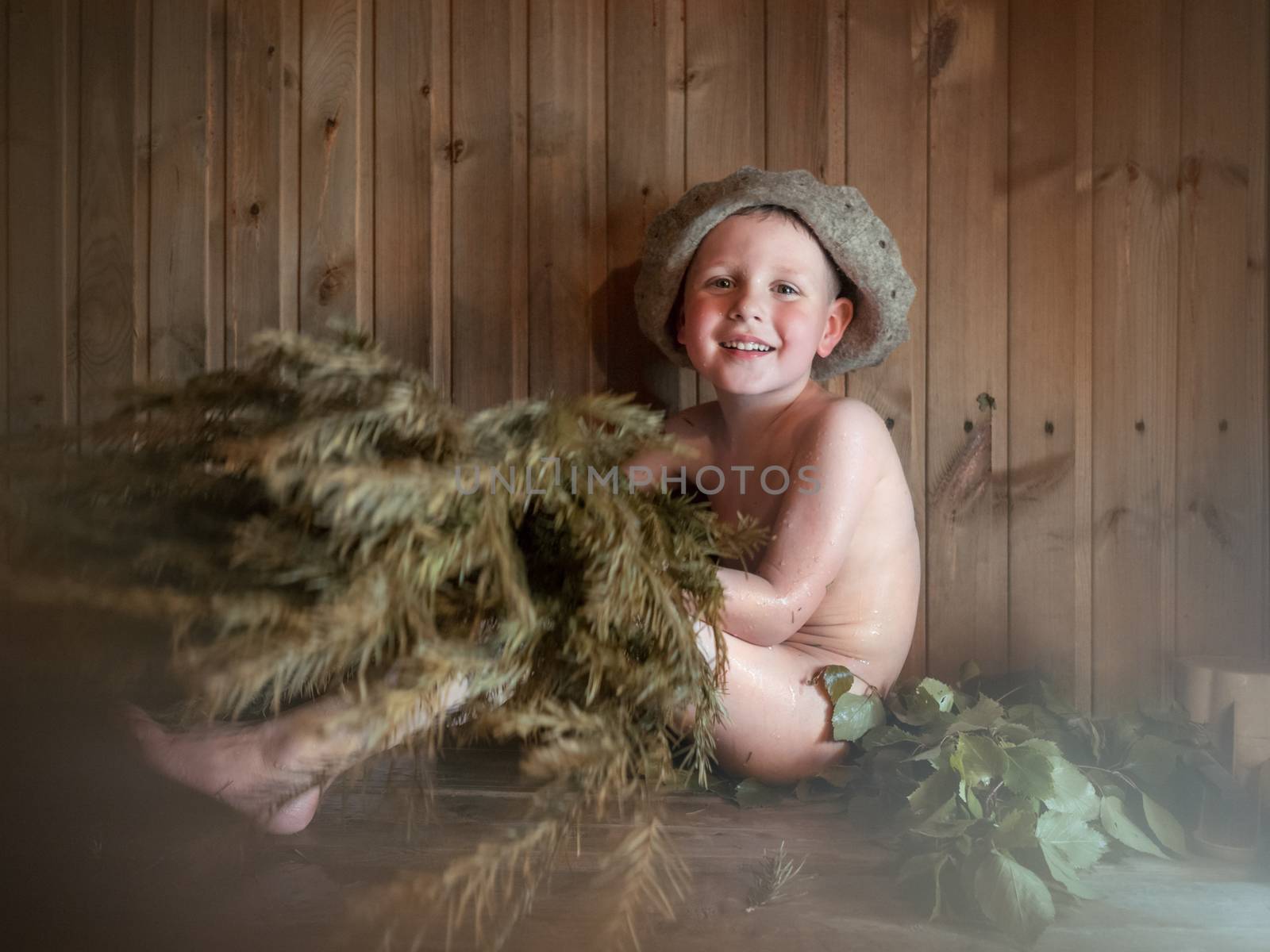 Four-year-old naked boy in hat with silver-fir broom in hands in Russian bath-house. Smiling little child is taking steam-bath at sauna. Authentic shot