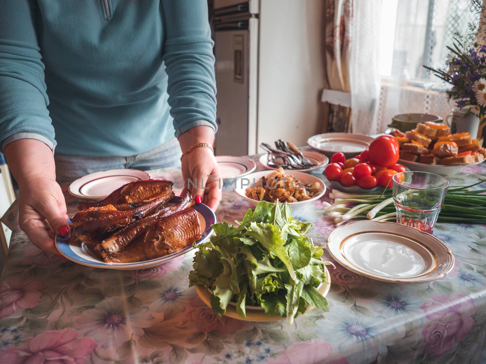 Woman sets kitchen table in country house. Woman hold plate with smoked fish on the table. Table setting in country house with wild flowers bouqet. Cottagecore and farmcore concept. Authentic shot