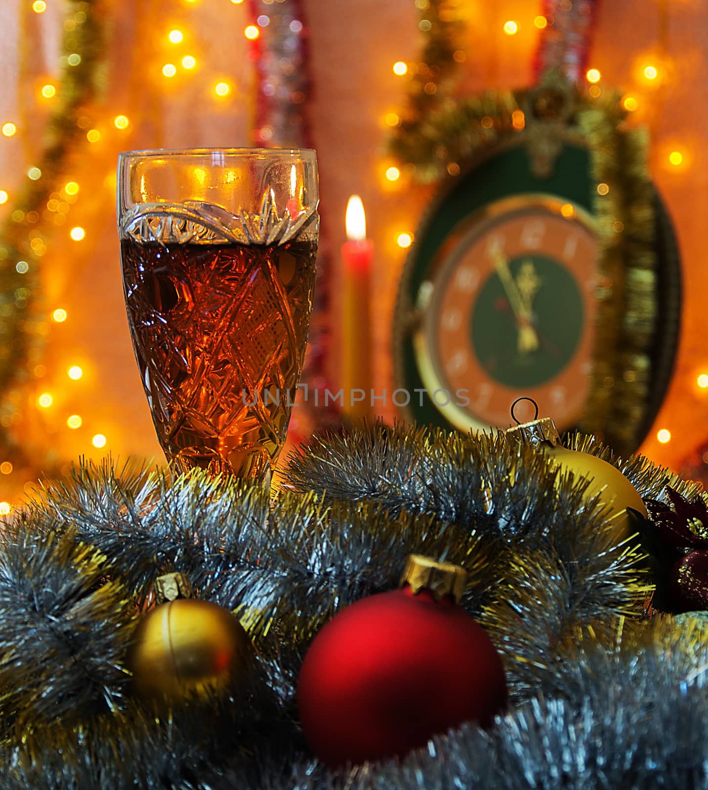 In the foreground is a wine glass with wine. Christmas ornaments lie around Fougeres. In the background is out of focus and see the candle clock, in which time five minutes to twelve. Background illuminated garland.

