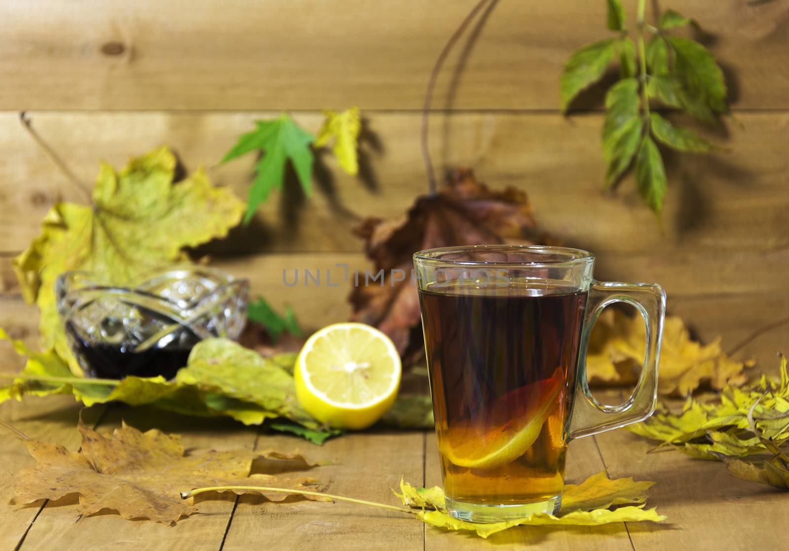 In the foreground is in focus glass cup of tea. In the background, the focus is visible without lemon, vase with jam and autumn leaves.