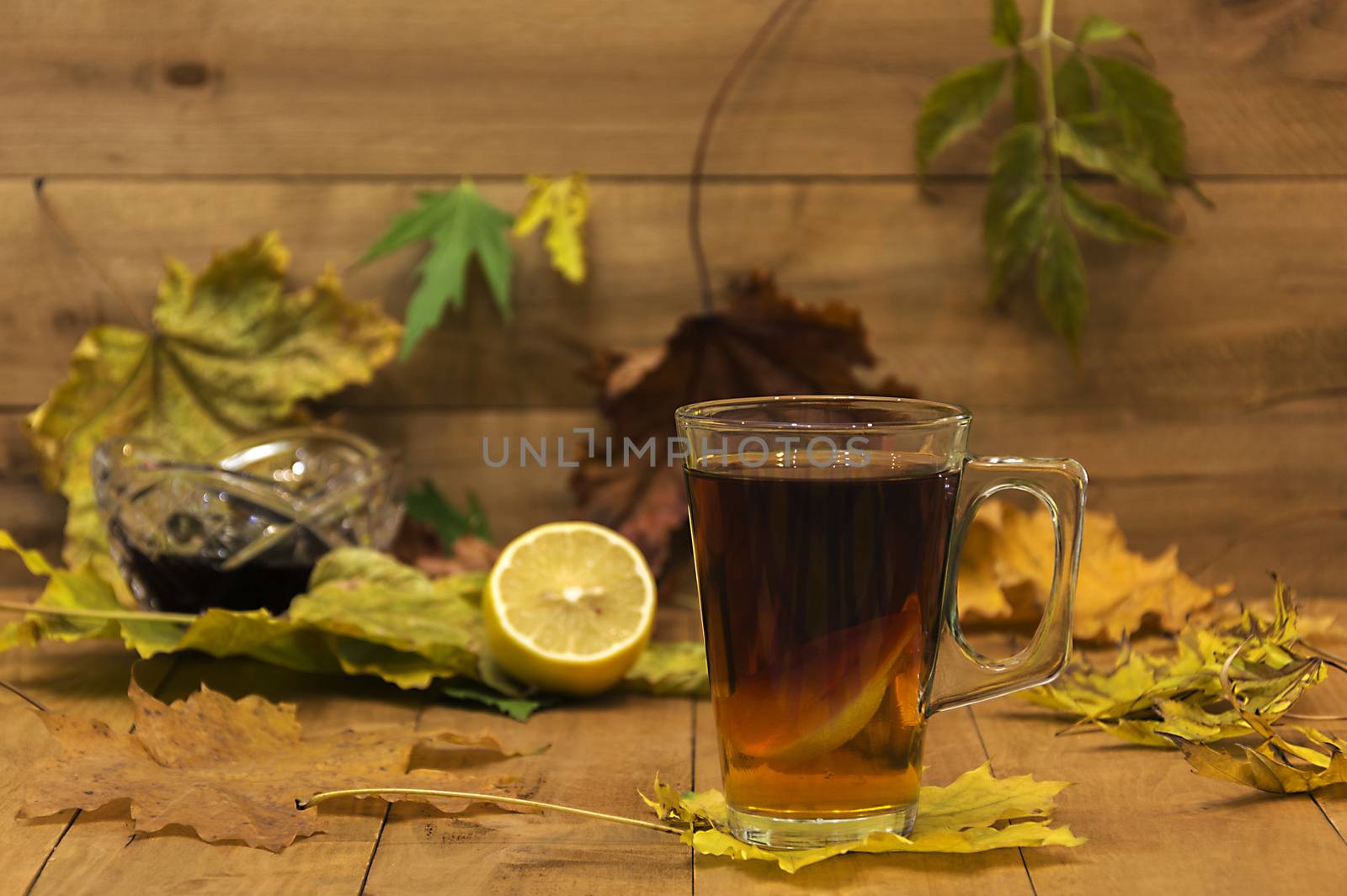 In the foreground is in focus glass cup of tea. In the background, the focus is visible without lemon, vase with jam and autumn leaves.