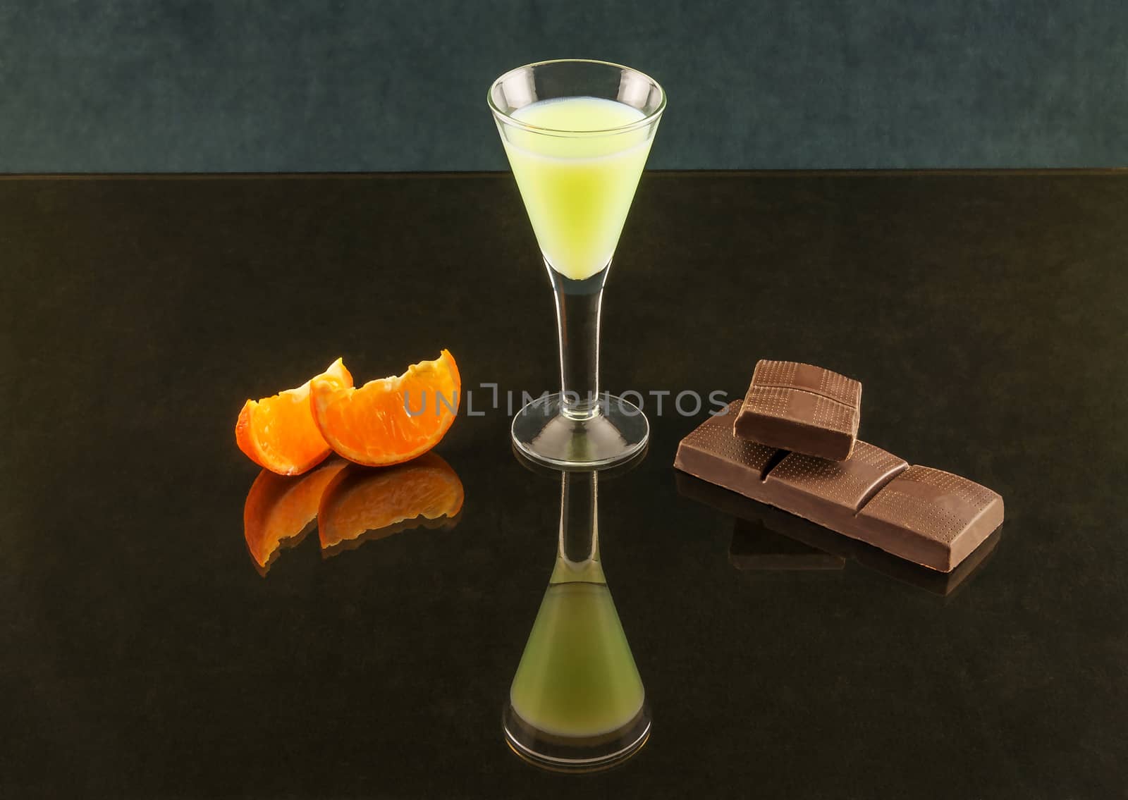 Wineglass on a thin stalk with a drink, chocolate and tangerine on a mirror surface with reflection